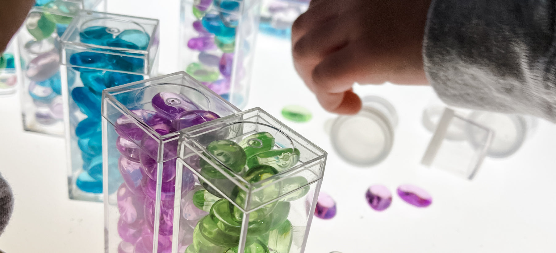 Colorful jewels in clear containers on a light table being played with by a young child