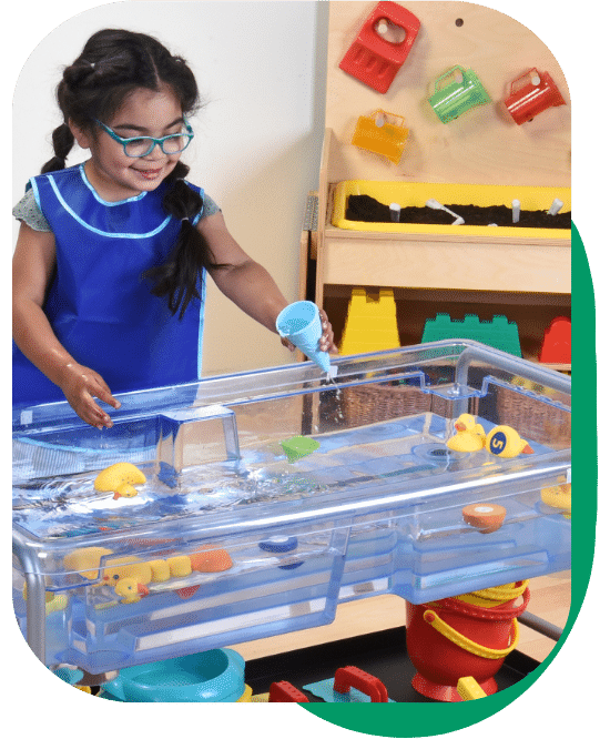 young girl wearing an apron playing with scoops and funnels at a water table in a classroom