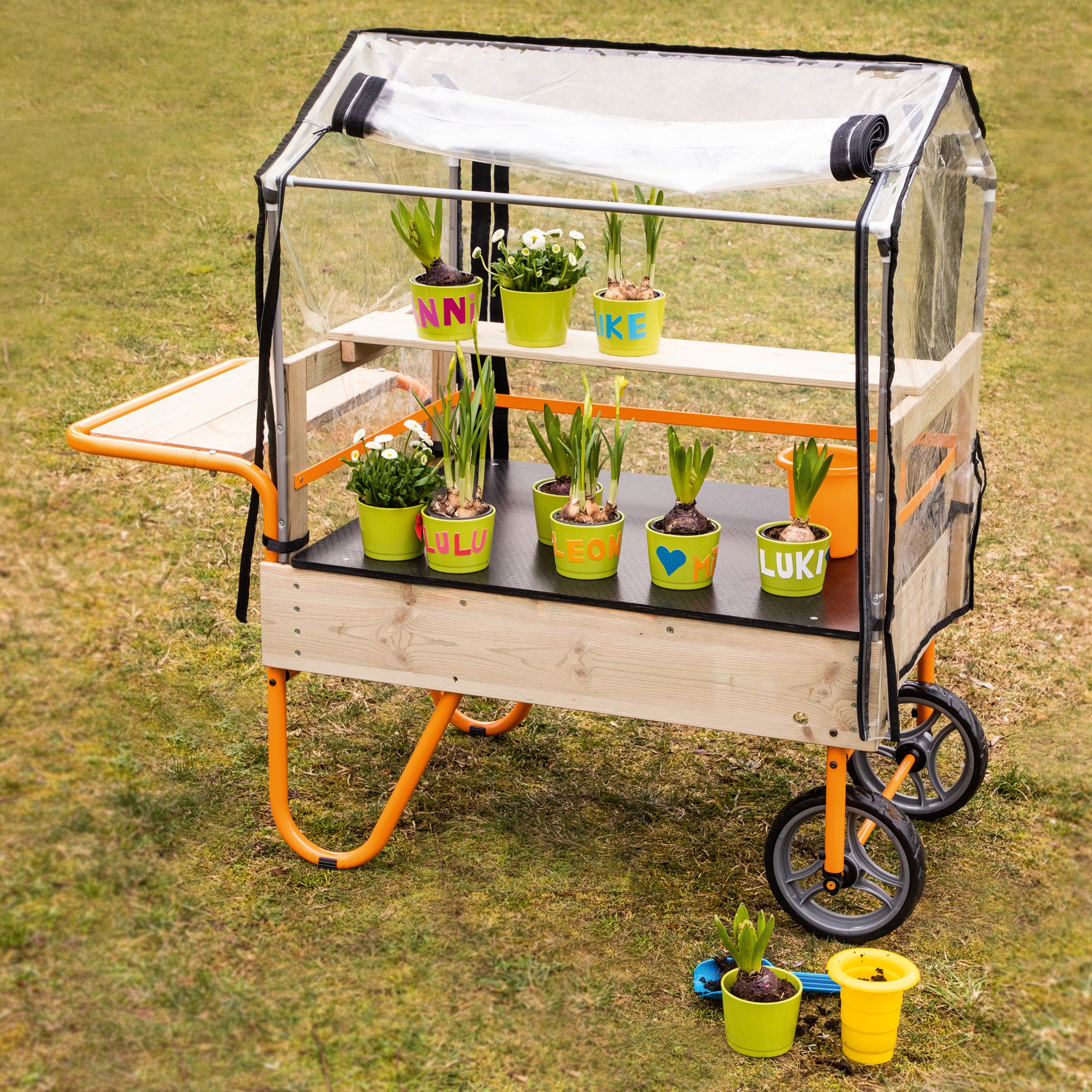A wooden three-tier garden cart with an attached clear plastic greenhouse dome and side tray, shown outdoors on grass with potted plants on the shelves and a few pots on the ground.