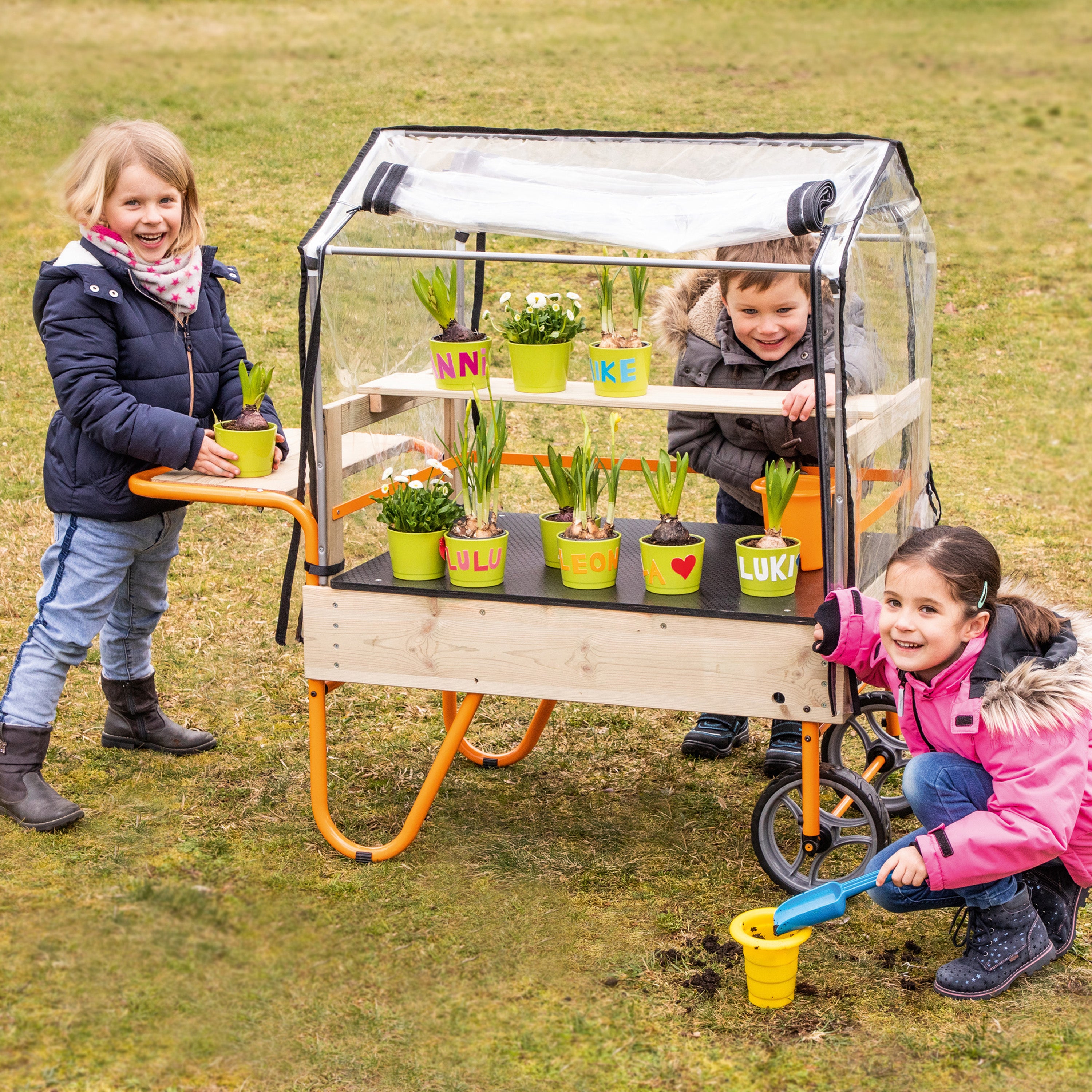 A children's wooden garden cart with a clear plastic greenhouse cover and orange metal legs, shown outdoors on grass with three children interacting with it and planting in small pots.