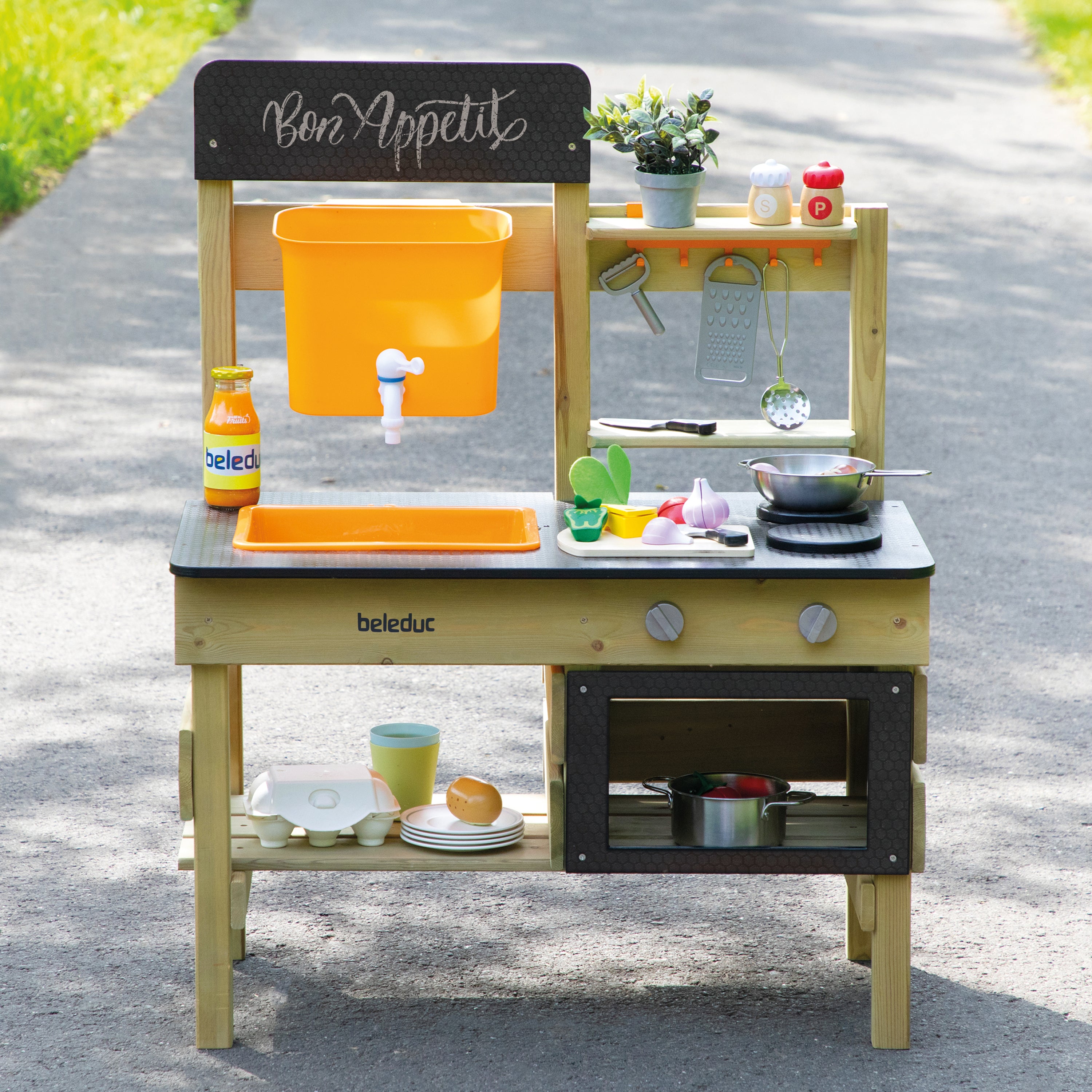 A wooden children's play kitchen set outdoors on a paved path. The play kitchen is made of light natural wood with black countertops and dark cabinet doors, featuring an orange hanging bin, a small sink, shelves with pretend food and utensils, and a small oven compartment.