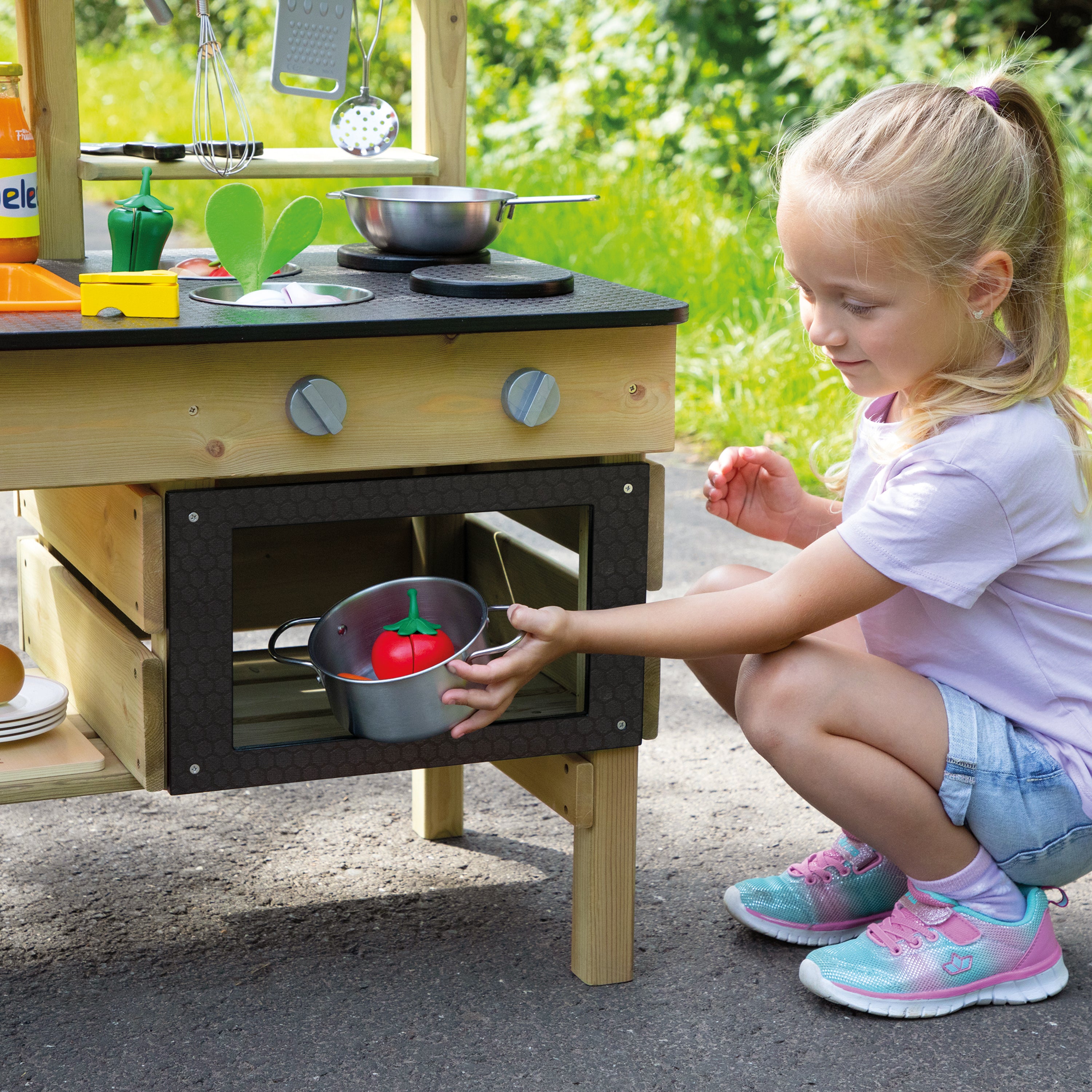 A young girl crouches beside a wooden outdoor play kitchen, reaching into an oven compartment where a small metal pot with a red toy pepper is sitting. The play kitchen has a light wood frame, a black countertop with pretend stove elements, and dark oven doors, set on pavement with grass in the background.