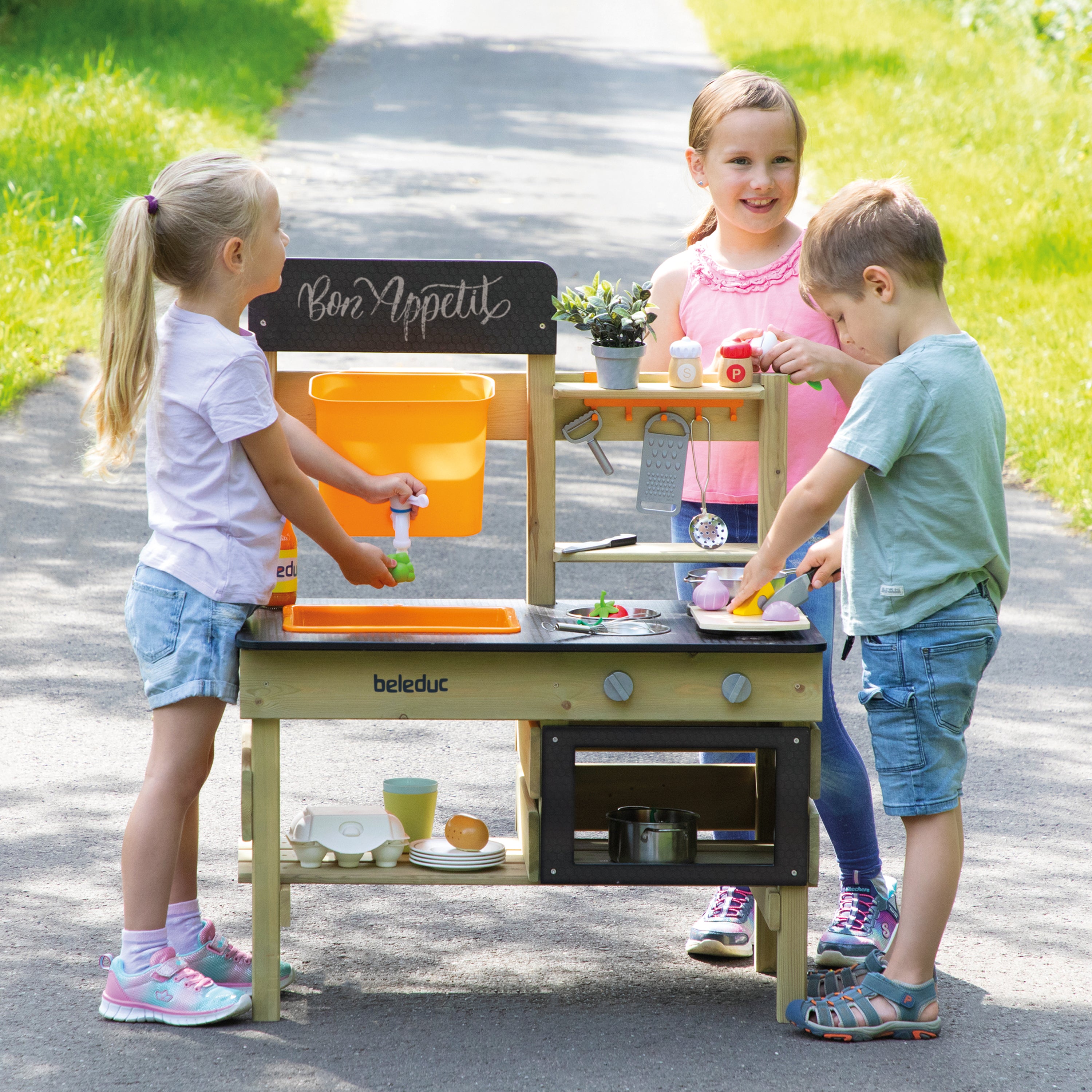 Three children playing with a wooden play kitchen outdoors on a paved path. The play kitchen has a light wood body, black front panel, and bright orange storage bins.