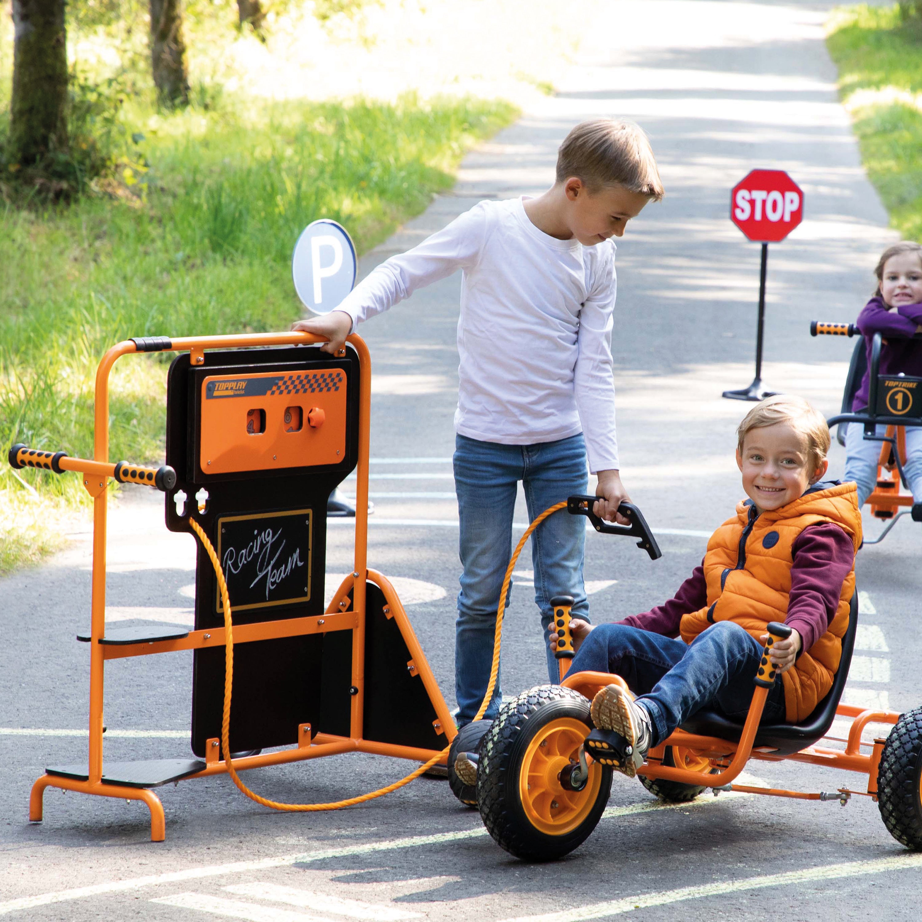 A child in a white shirt pushes an orange and black electric tricycle with a charging station attachment on a paved path, while another child sits in the tricycle. A second child is visible on a tricycle in the background.
