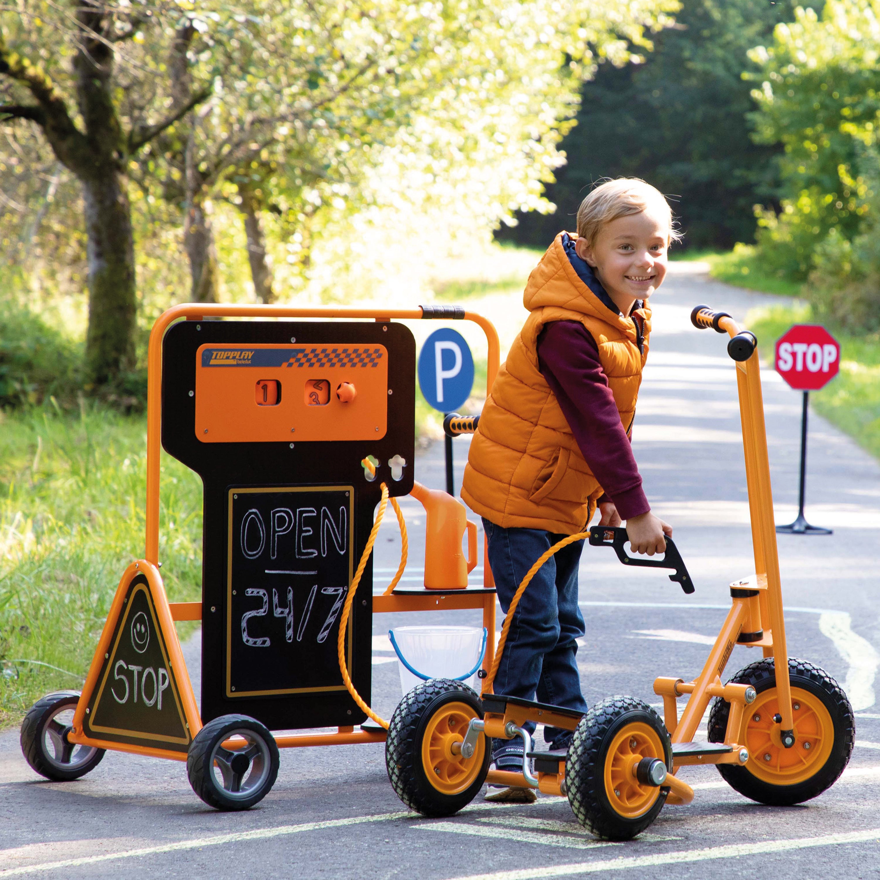 A child wearing an orange puffer vest rides an orange and black toy vehicle with training wheels on a paved path outdoors. The toy has a chalkboard-style front panel, a stop sign on the front, and a fuel cap accessory attached.