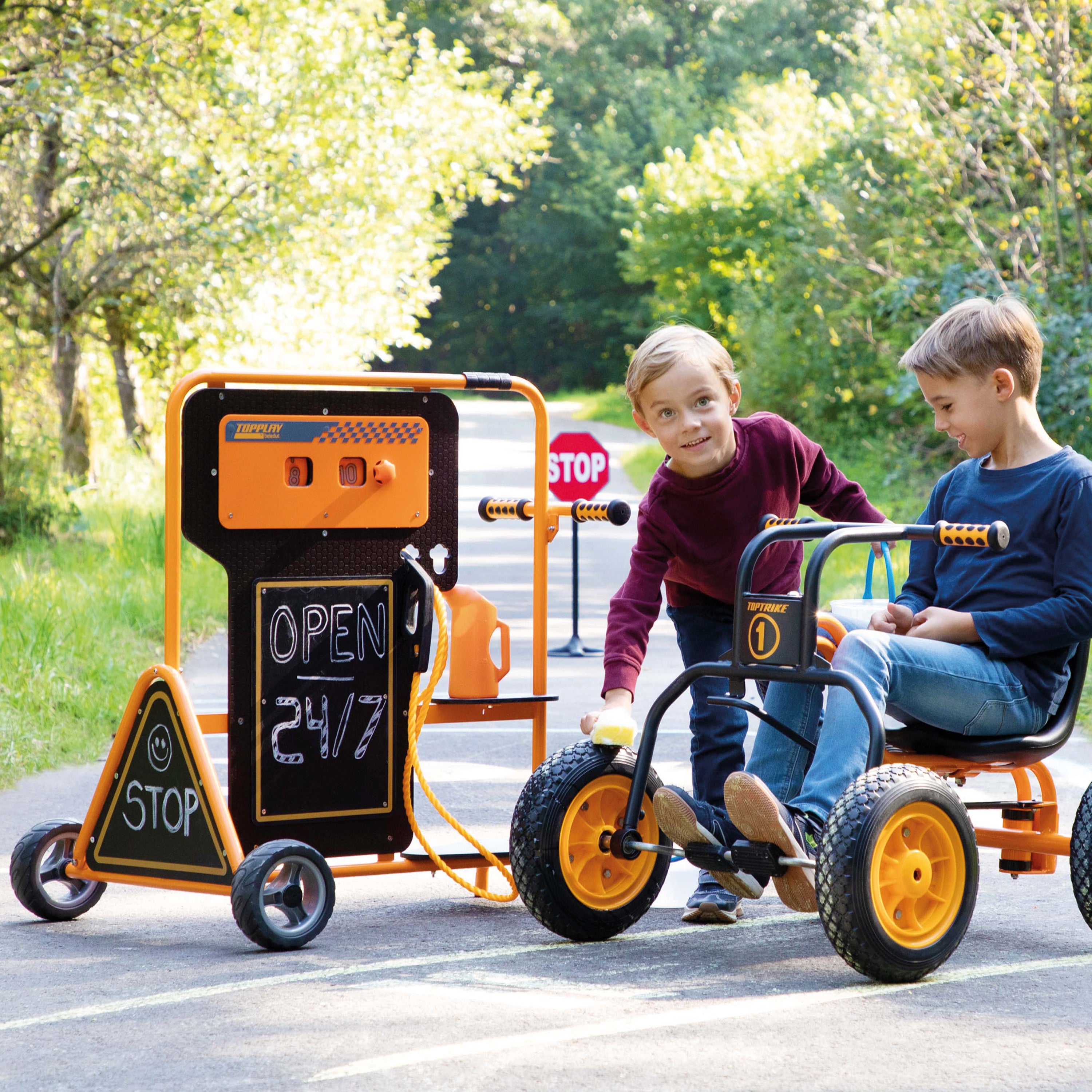 Two children outdoors on a paved path next to an orange and black utility cart with chalkboard signs and a small tricycle-style cart with yellow wheels.