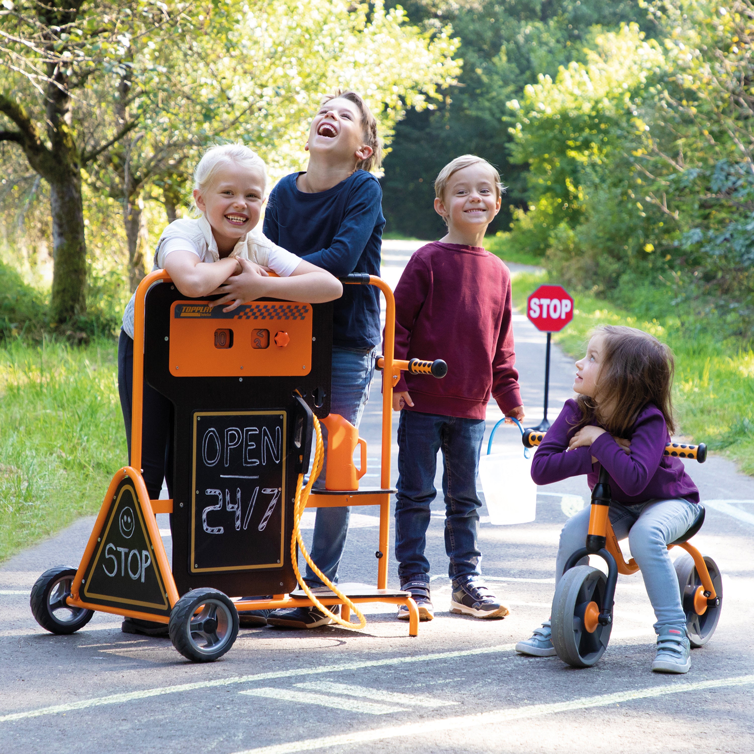 Four children outdoors on a paved path near green trees, interacting with an orange and black playground cart. One child is leaning on the cart, one is laughing, one is standing nearby, and one is sitting on a tricycle beside the cart.