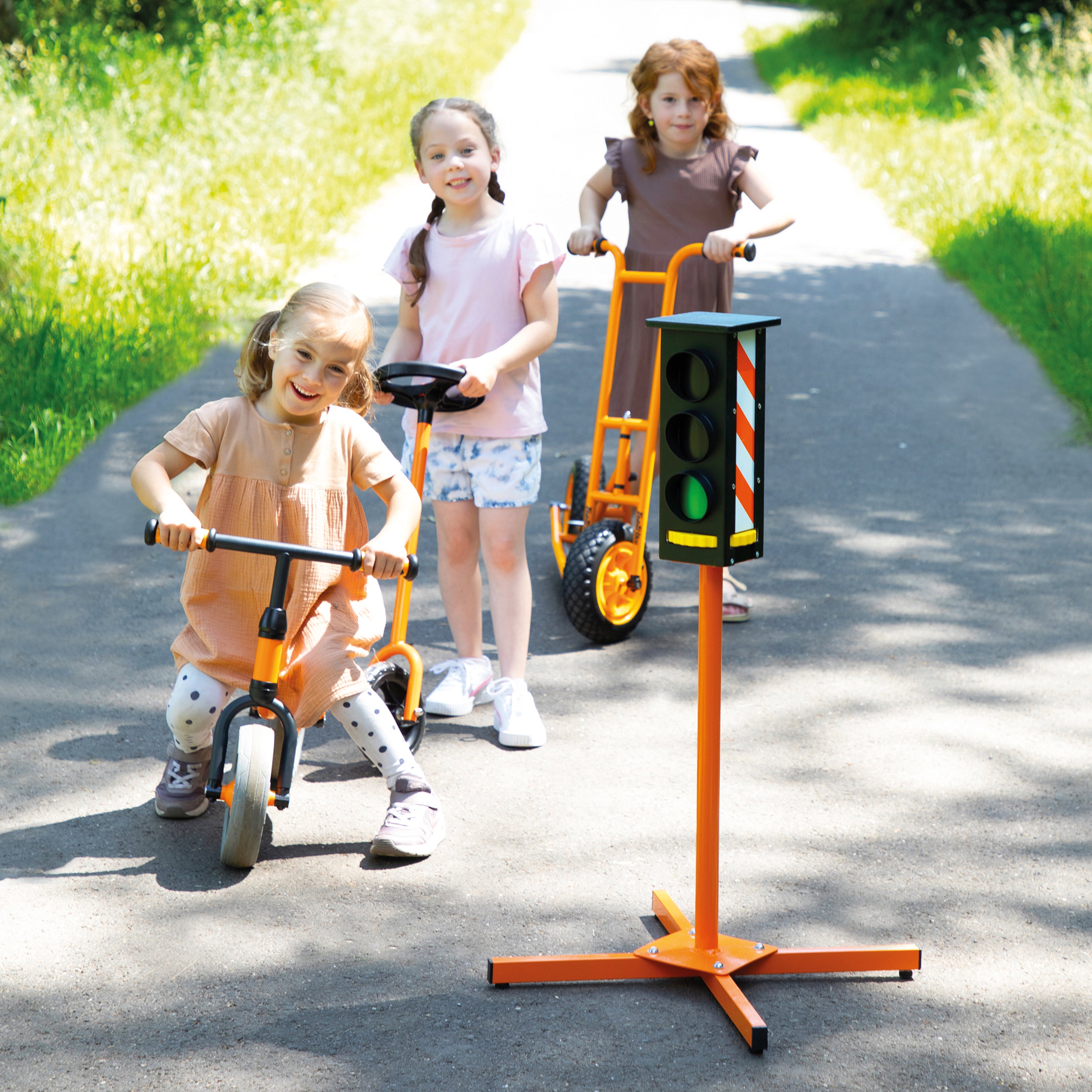 Three children outdoors on a paved path using orange toy vehicles, with one child in the foreground riding an orange balance bike and another standing near a toy traffic light on a stand.