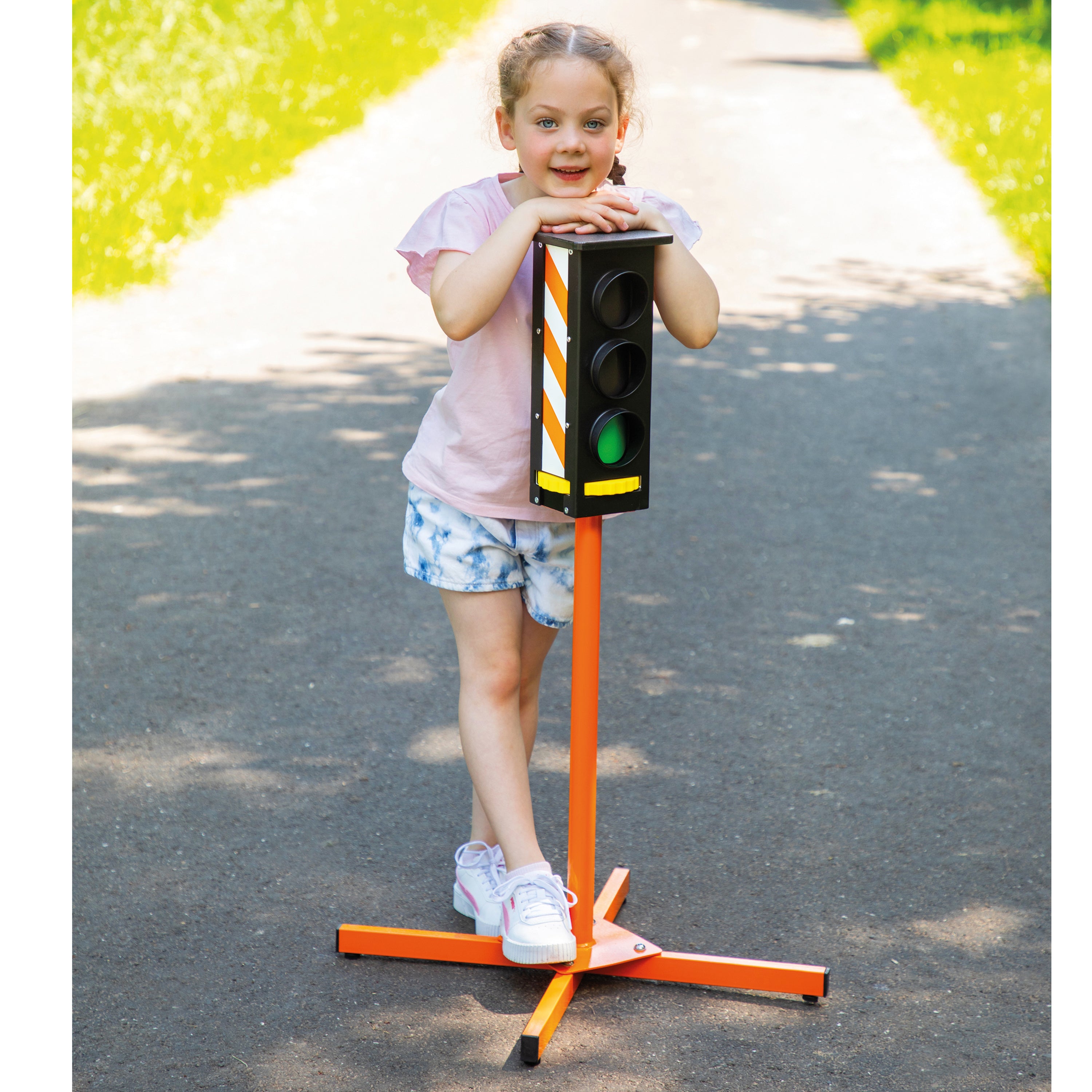 A young girl stands on an orange traffic light stand in an outdoor park setting, holding the black and yellow signal head. The product being shown is the portable traffic light toy/sign.