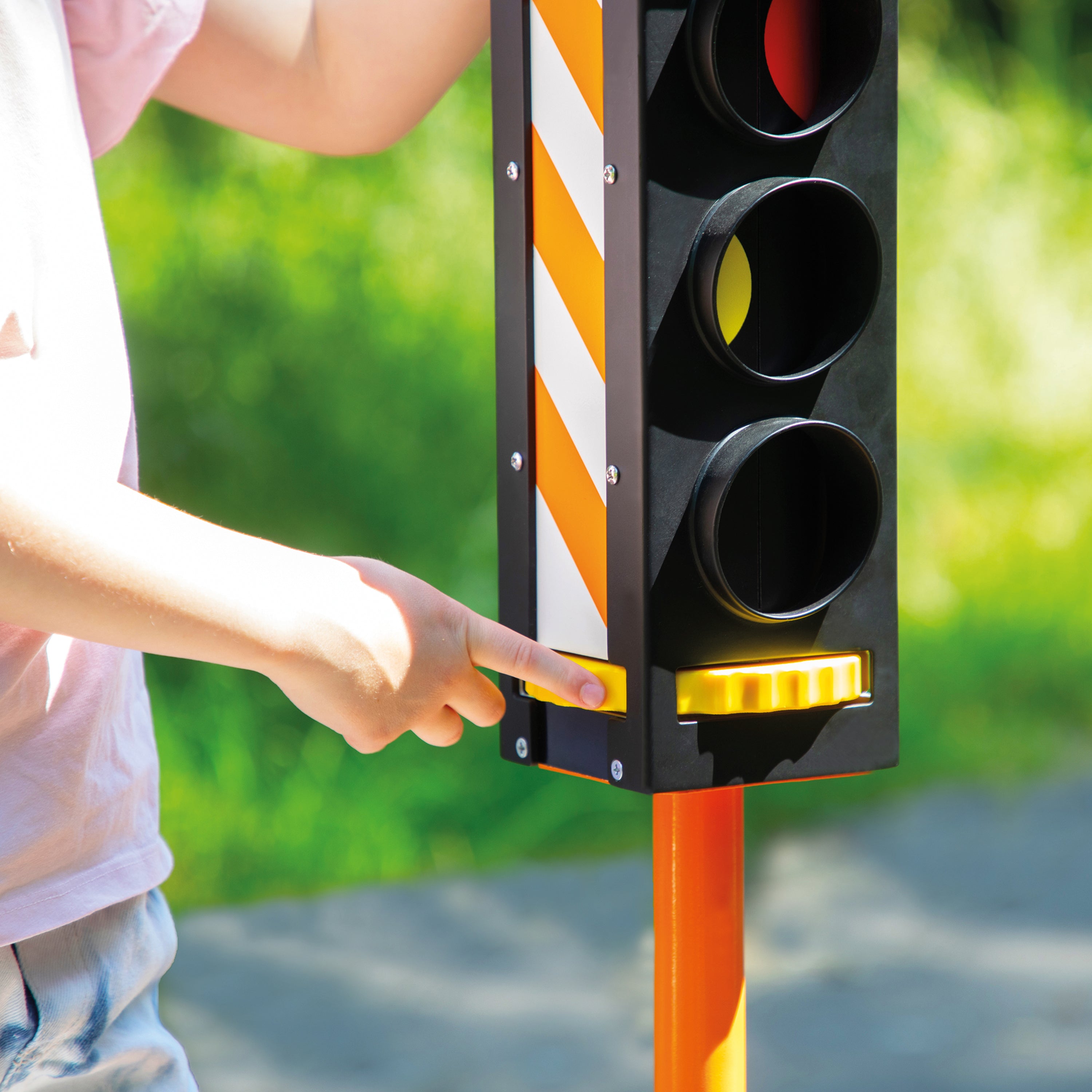 A close-up of a child's hand pressing a yellow button on a traffic light play structure, with a blurred green outdoor background.