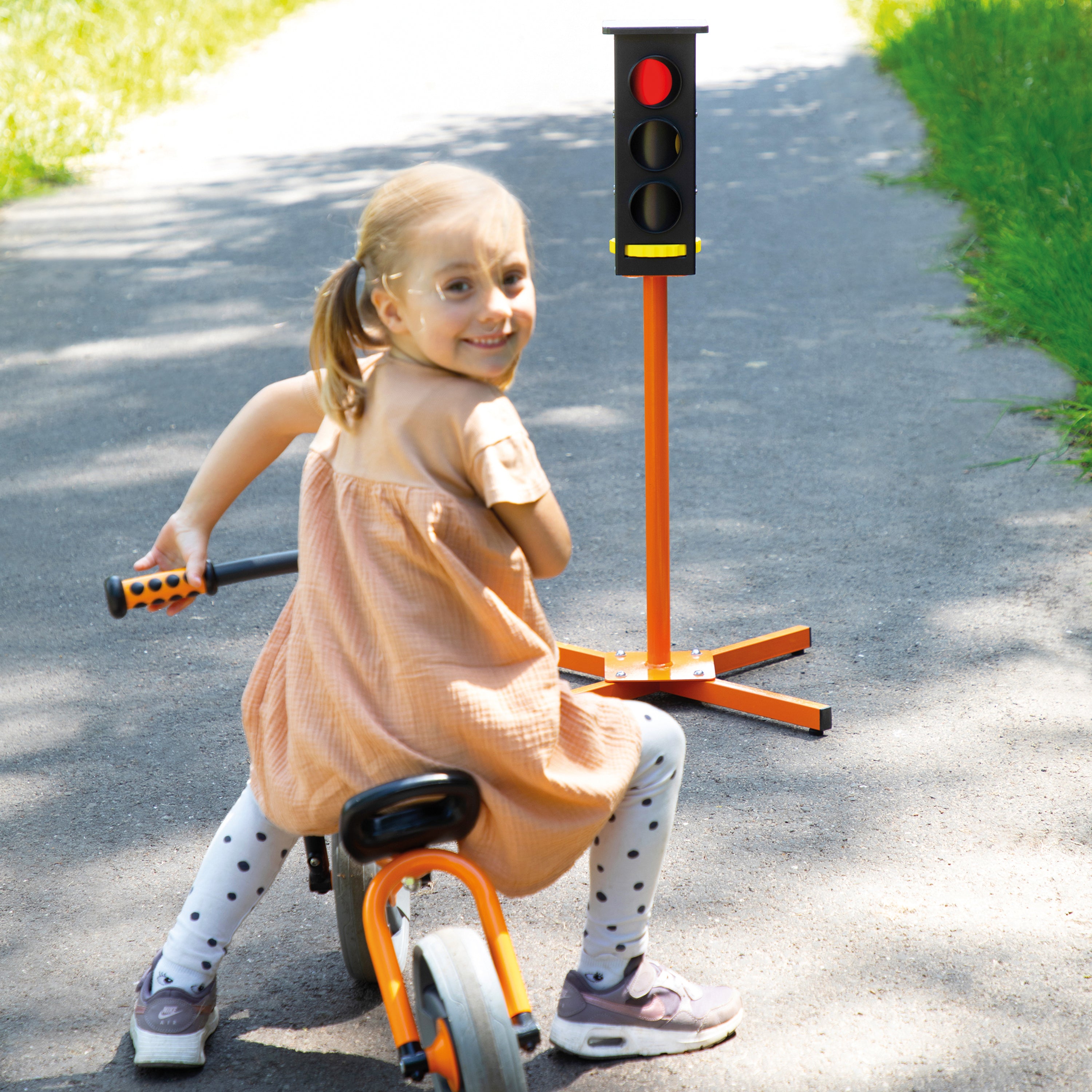 A young girl riding a balance bike outdoors on a paved path, holding a handlebar while facing a traffic light toy mounted on a stand. She is wearing a peach dress, white leggings with black polka dots, and gray sneakers.