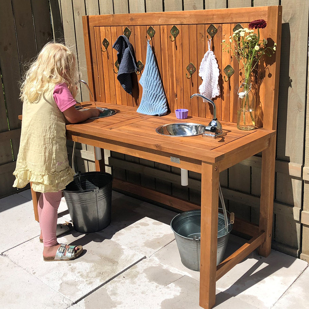 Child playing at an outdoor wooden play sink with various items on a sunny day.