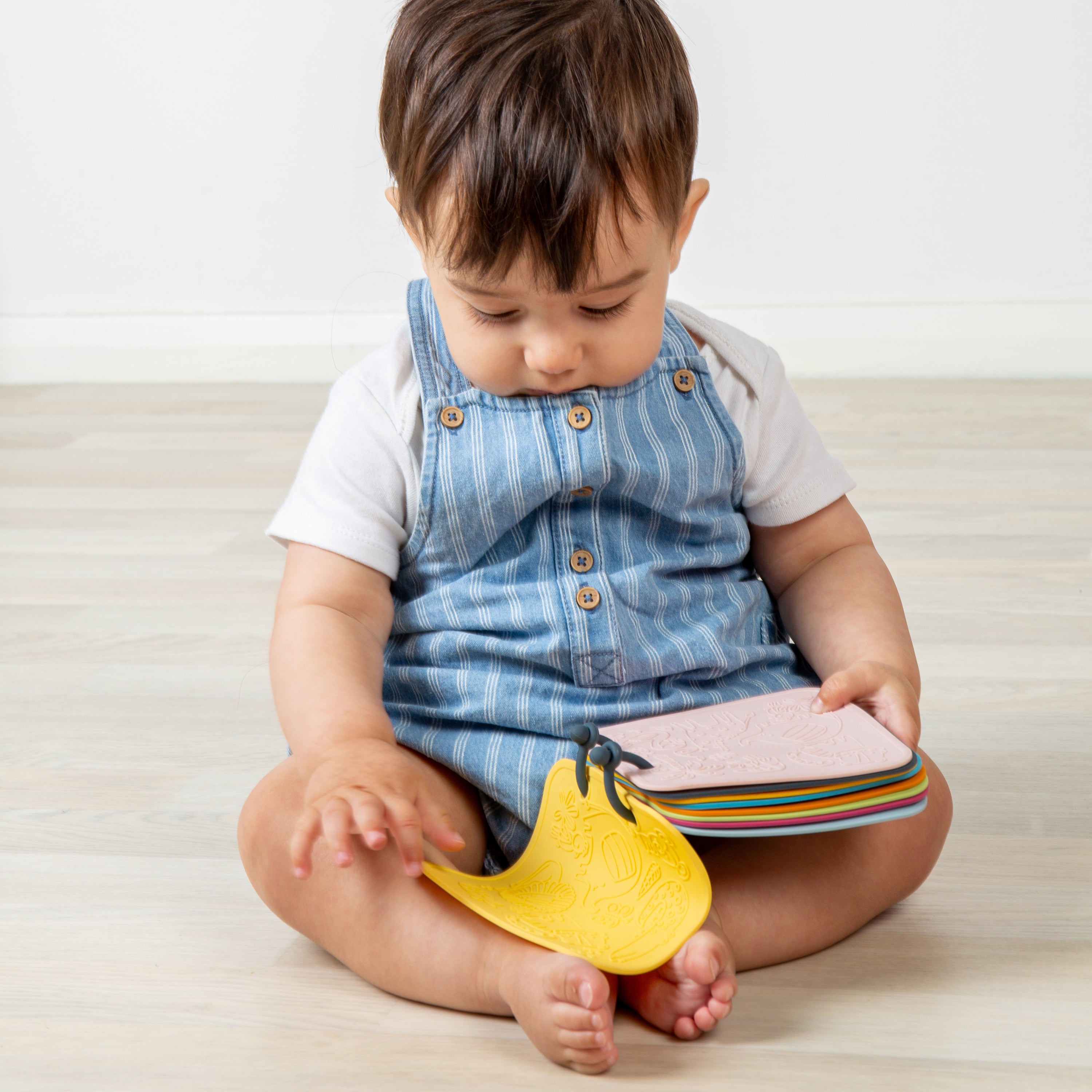 Child sitting on a light-colored floor holding a colorful book and a yellow toy.