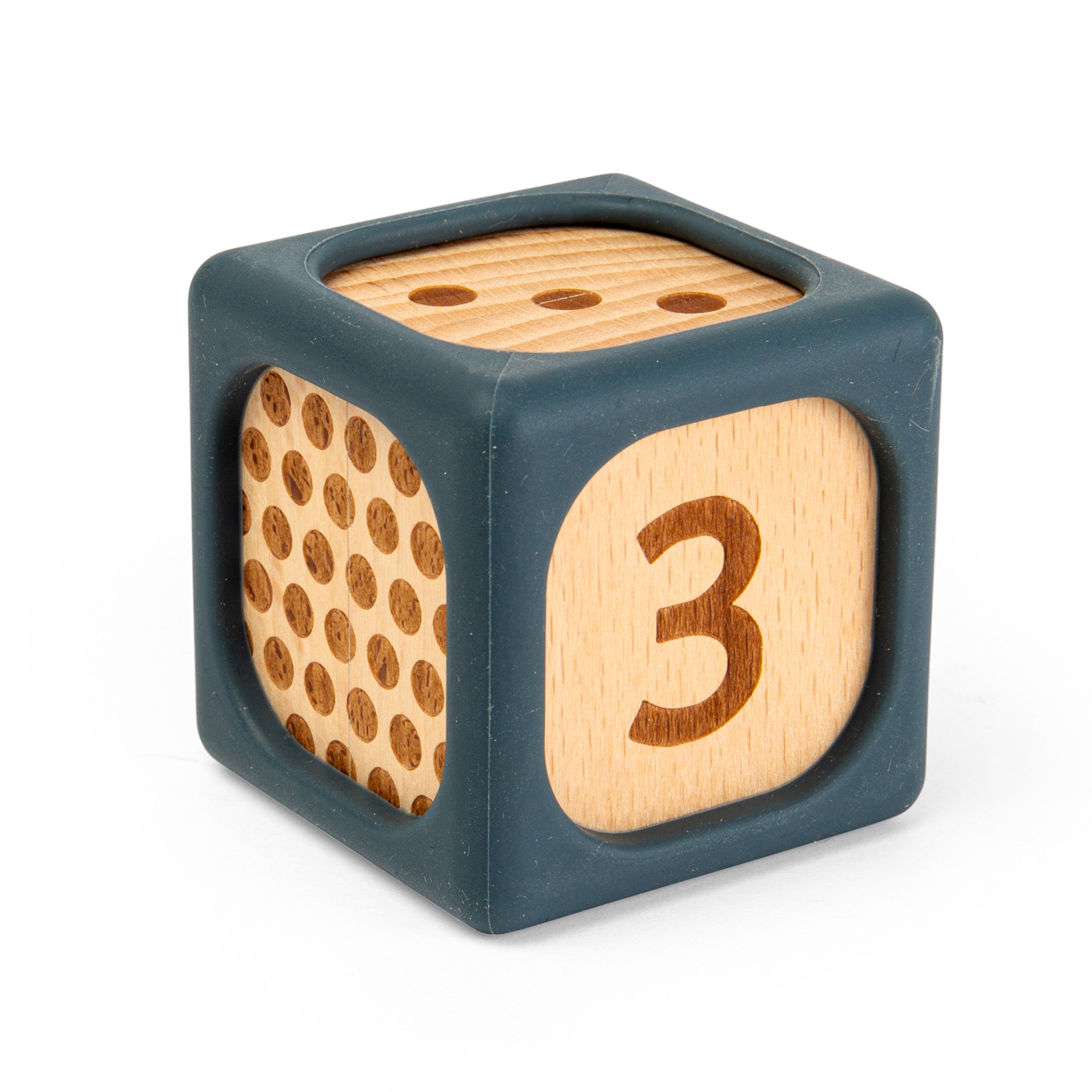 A wooden toy cube with a dark blue-gray silicone outer frame. One side shows a natural wood panel with brown circular dots, and another side shows a natural wood panel with the number 3 printed in brown.