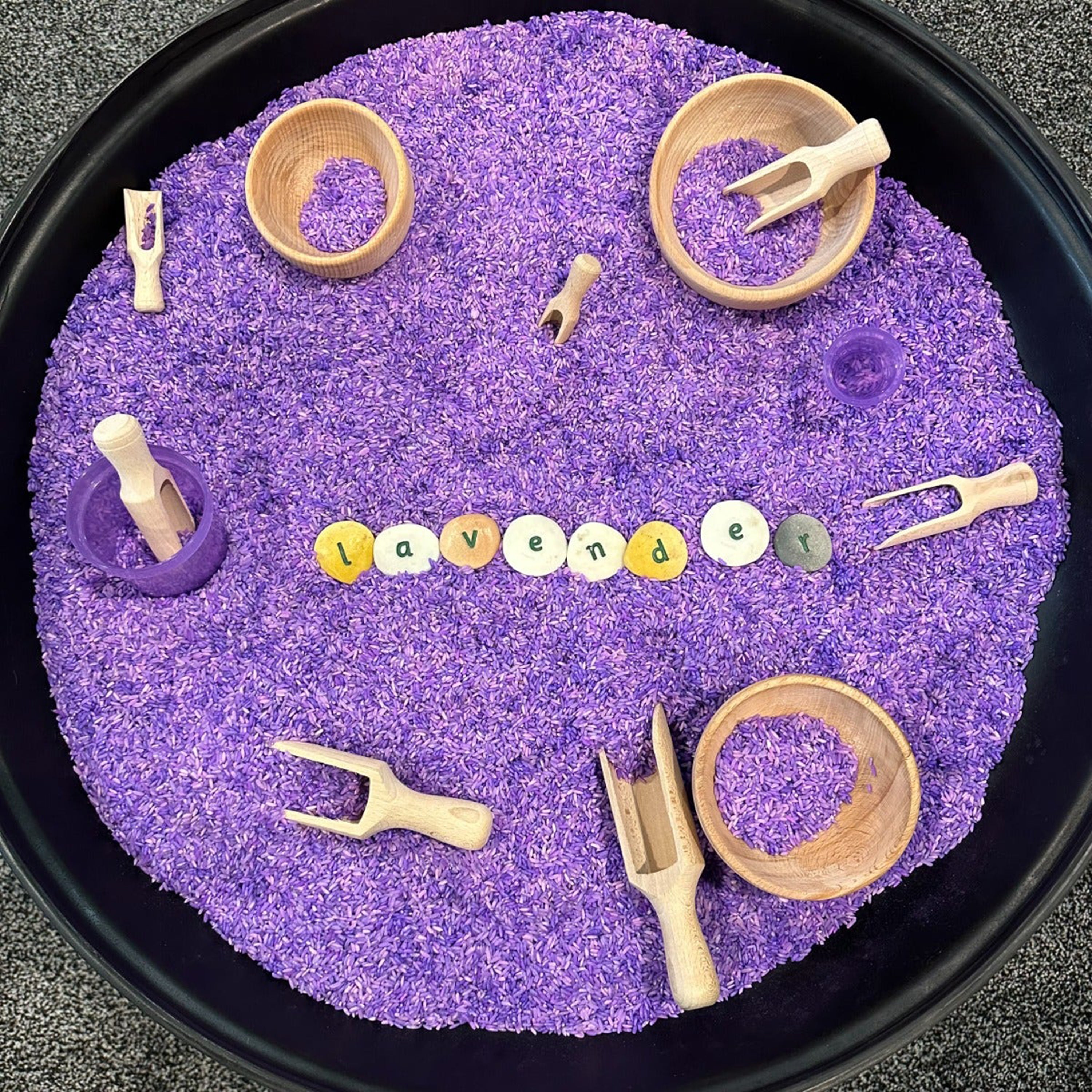 Purple sand with wooden tools and colorful letters spelling 'Lavender' on a black tray.