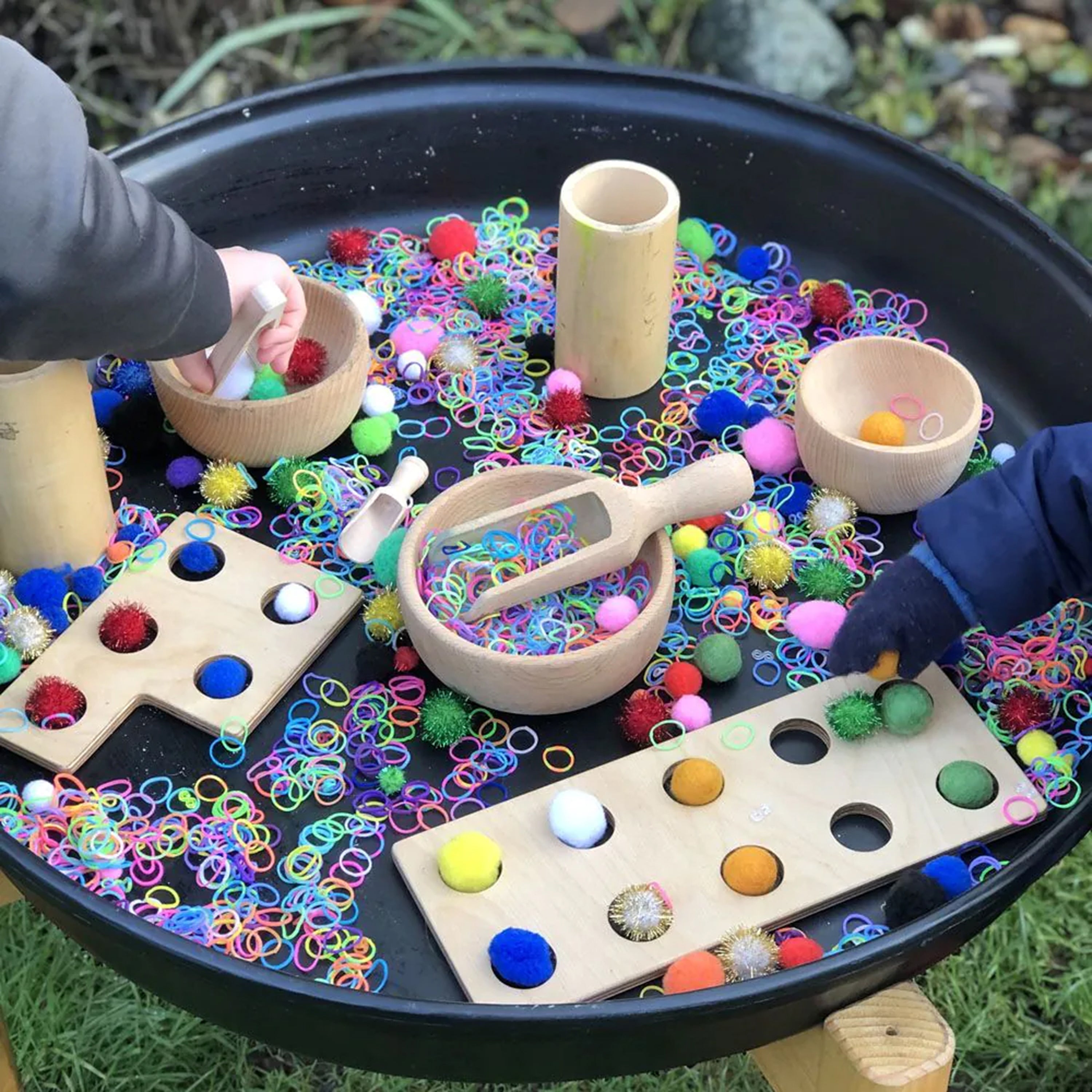 Colorful sensory play setup with beads, balls, and wooden toys on a black tray outdoors.