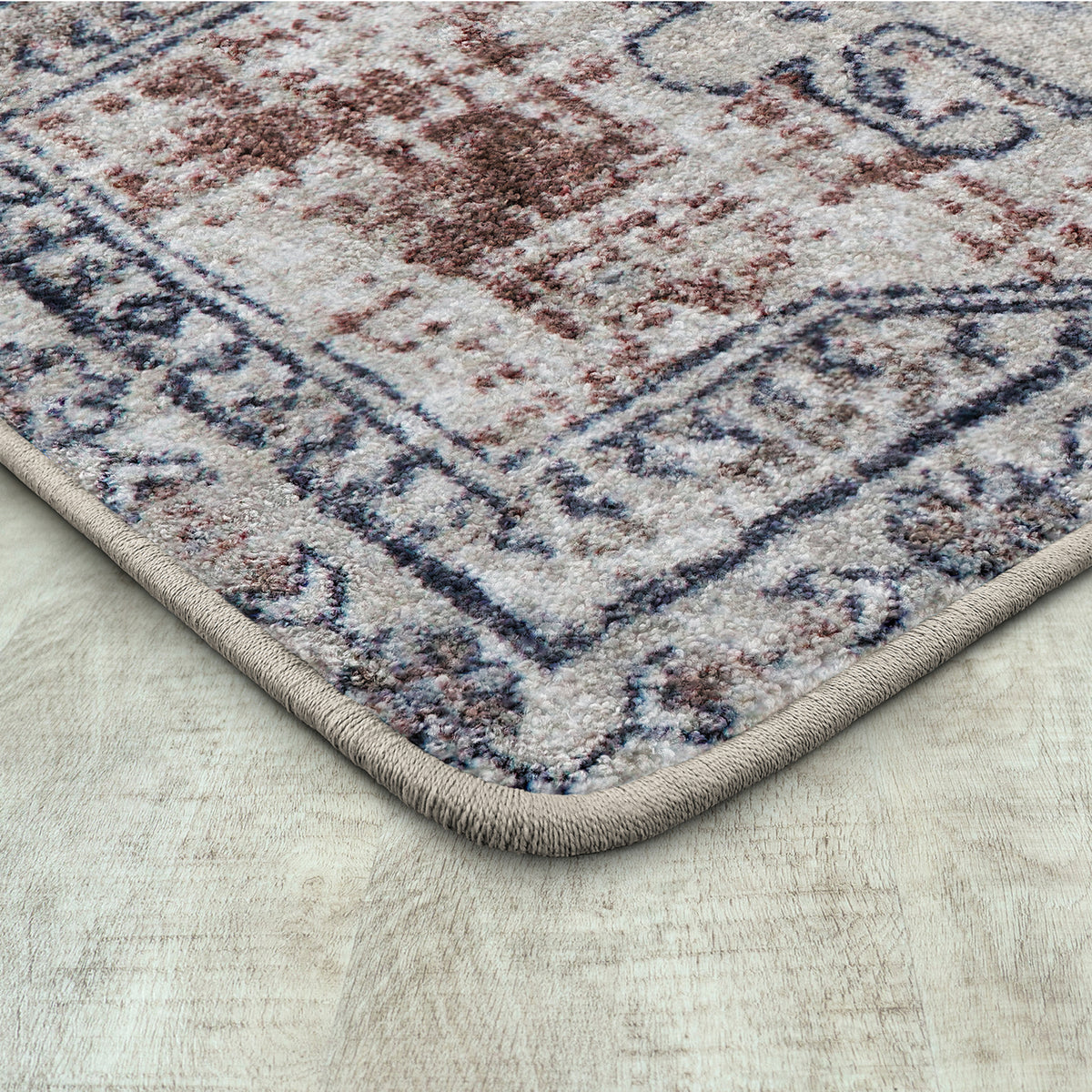 A close-up of a traditional area rug corner showing its beige and brown medallion pattern with a navy blue border, placed on a light wood floor.