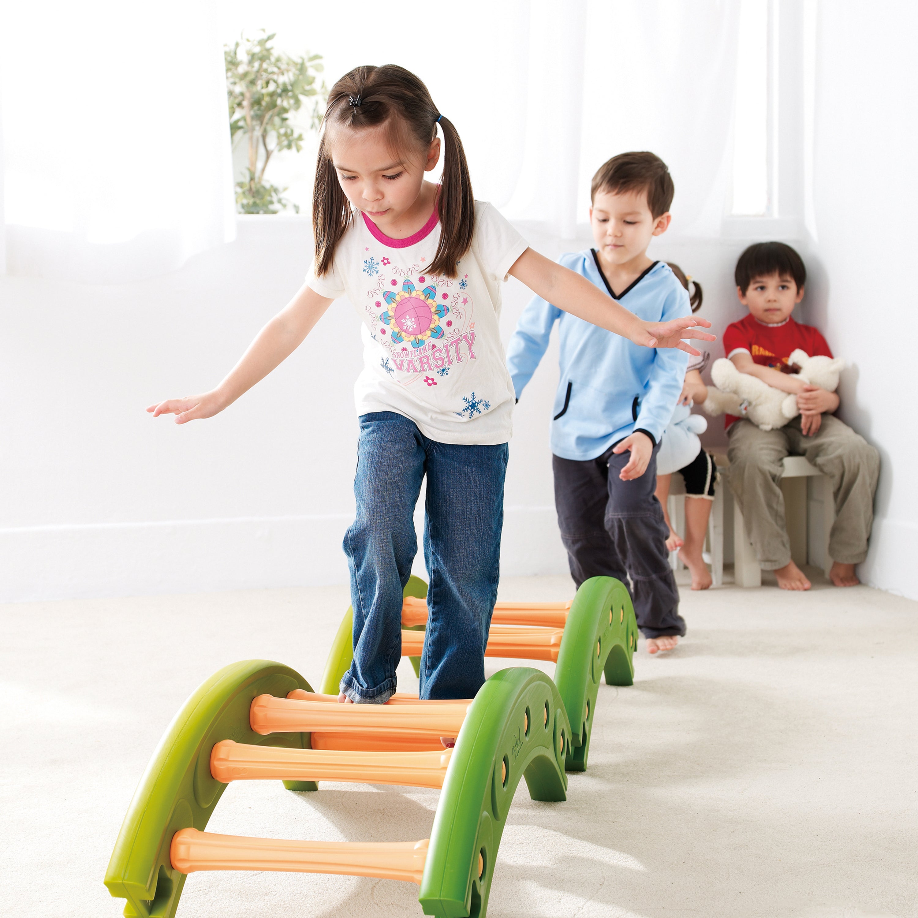 Children playing with a colorful balance beam on a white floor.