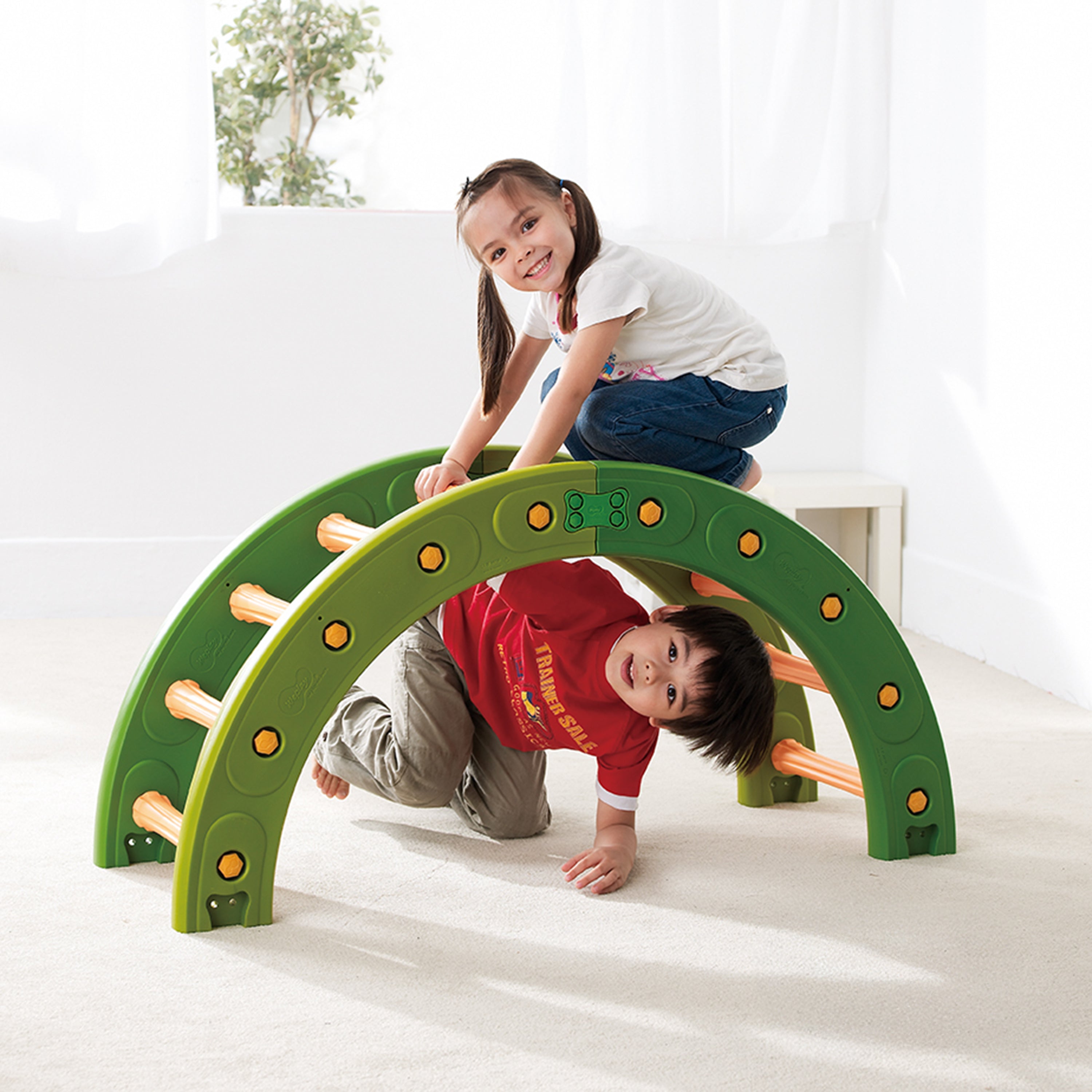 Two children playing on a green arch-shaped play structure indoors.