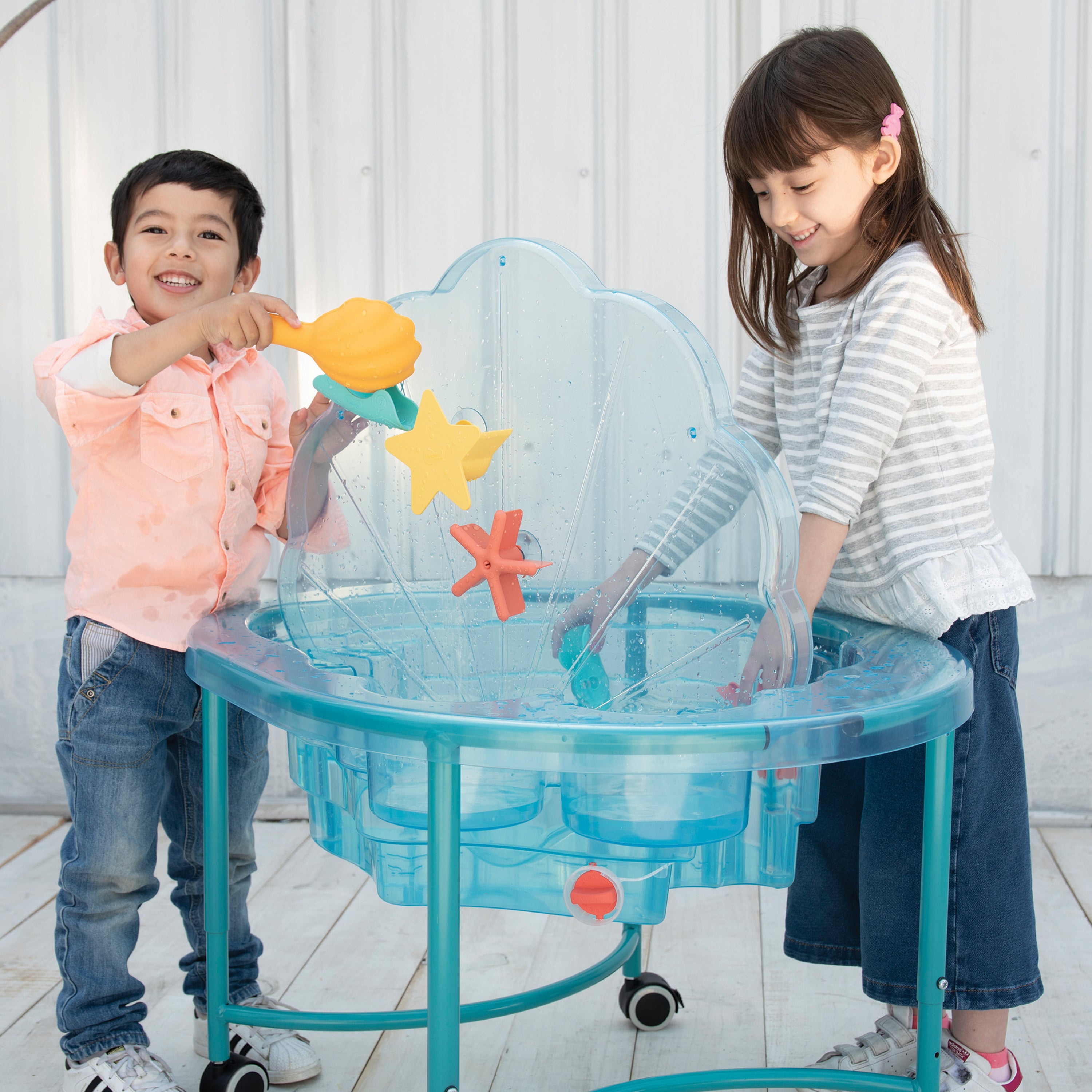 Two children playing with a transparent blue sensory table filled with water toys on a light wooden floor.