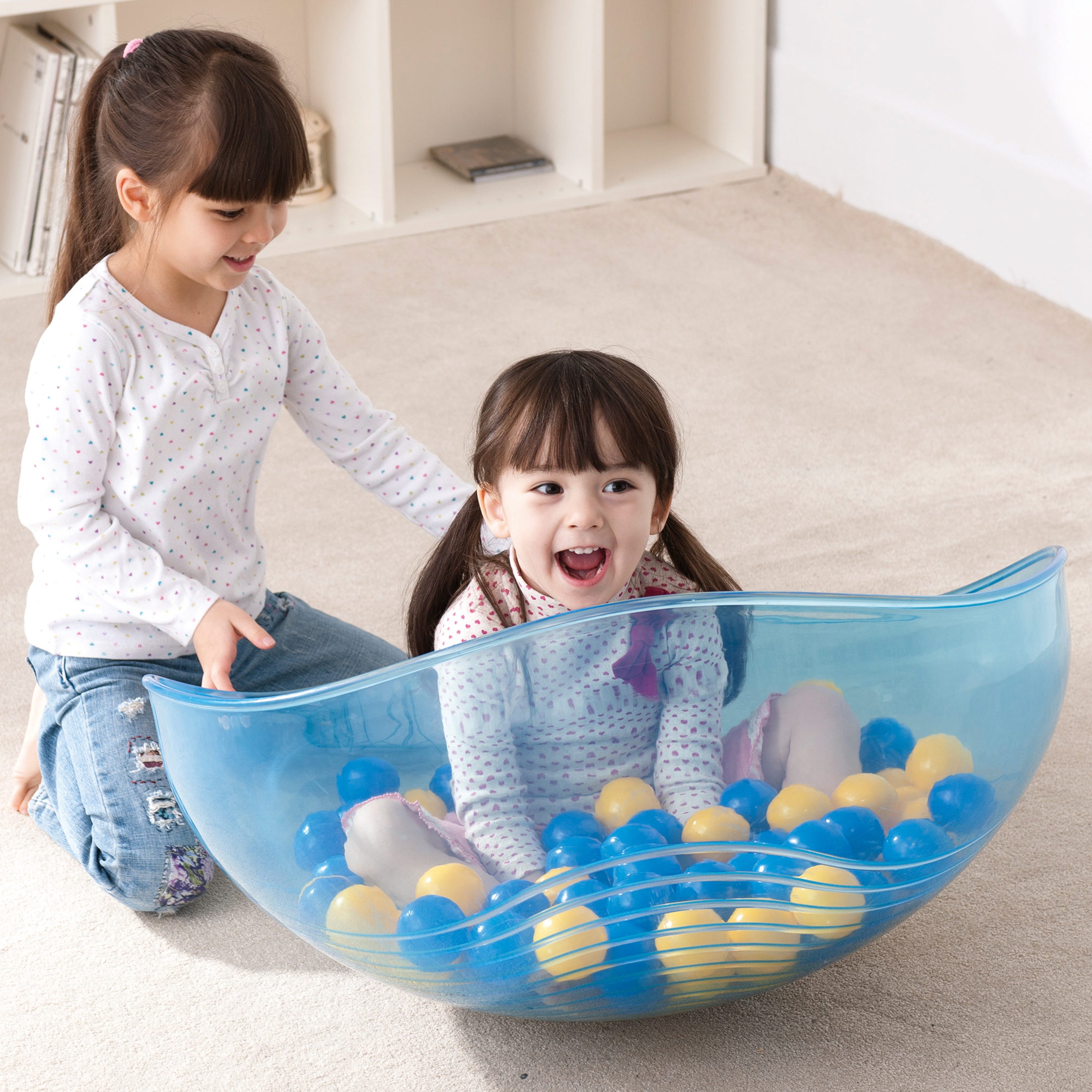 Two children playing with a large blue ball pit filled with colorful balls on a light-colored floor.