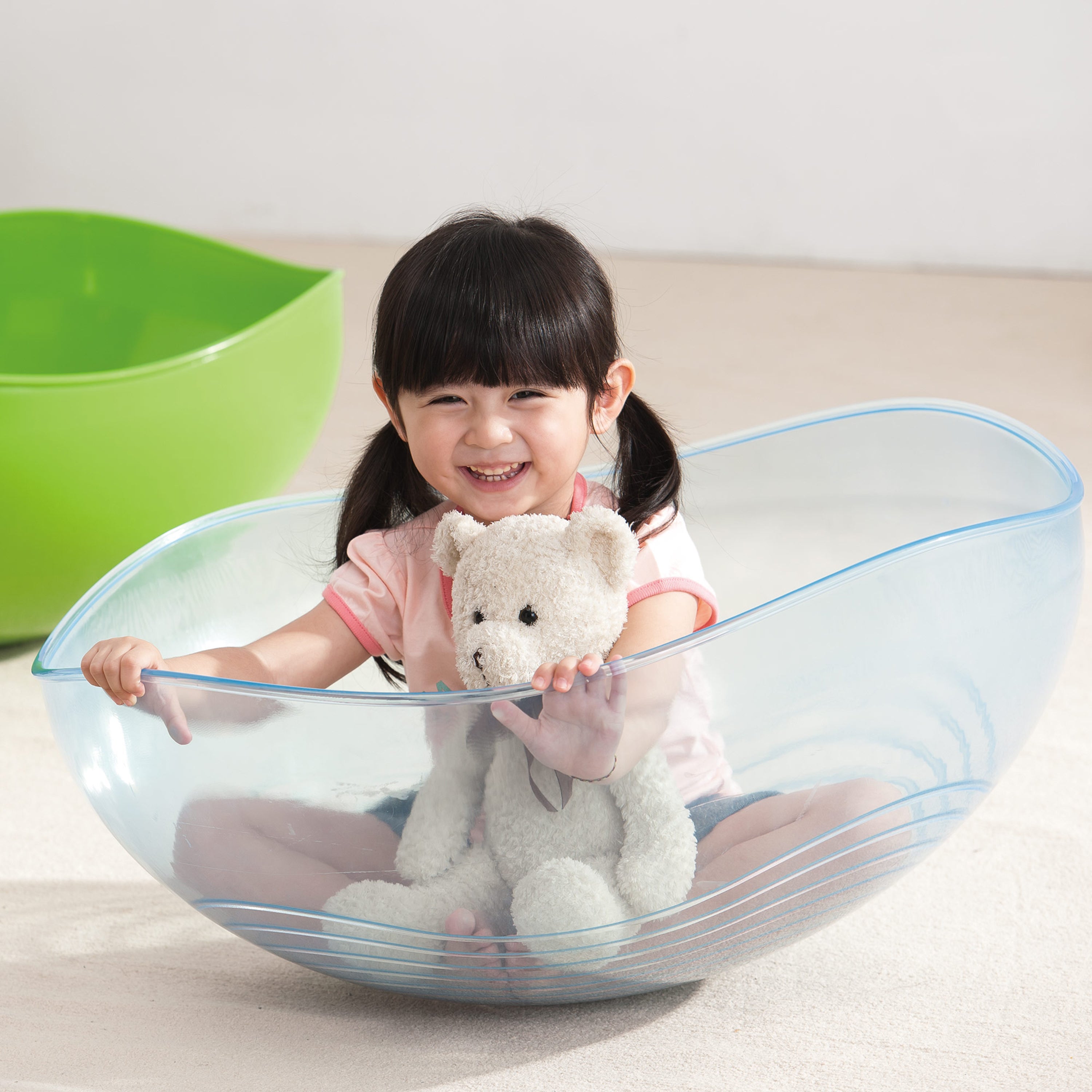Child holding a teddy bear inside a clear plastic chair with a green chair in the background.