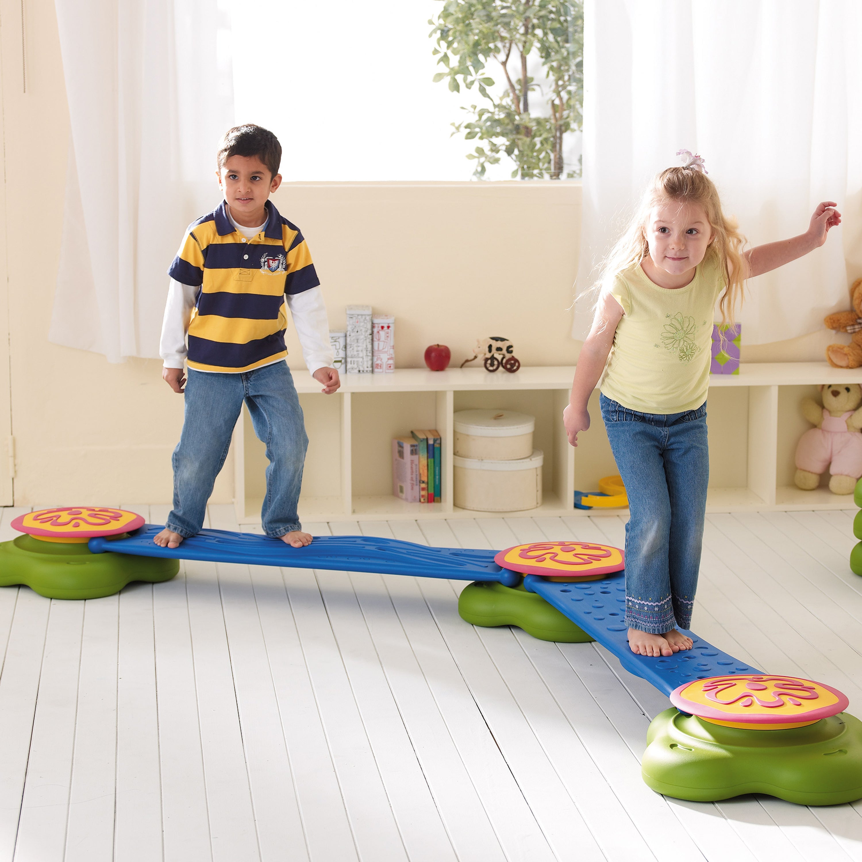 Two children playing on a balance beam toy in a room with a white curtain and shelves.