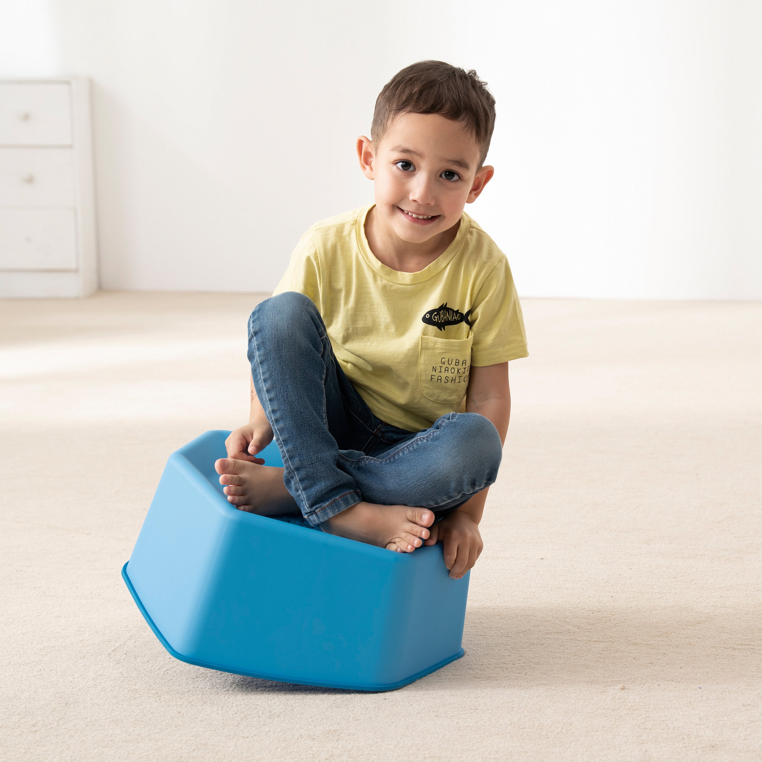 Child sitting on a blue stepping stone in a room with a white wall and beige carpet.