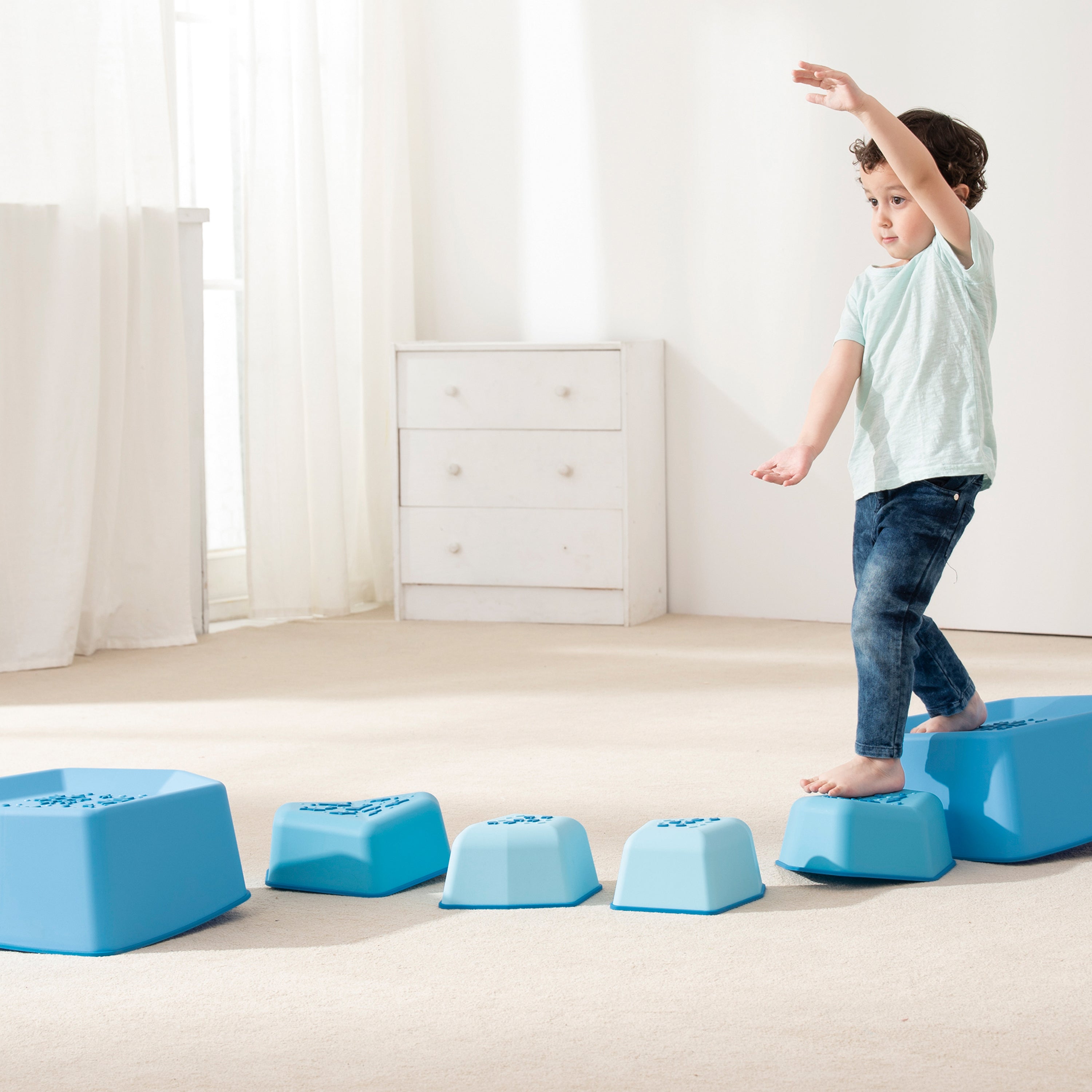 Child balancing on blue foam steps in a room with white curtains and furniture.