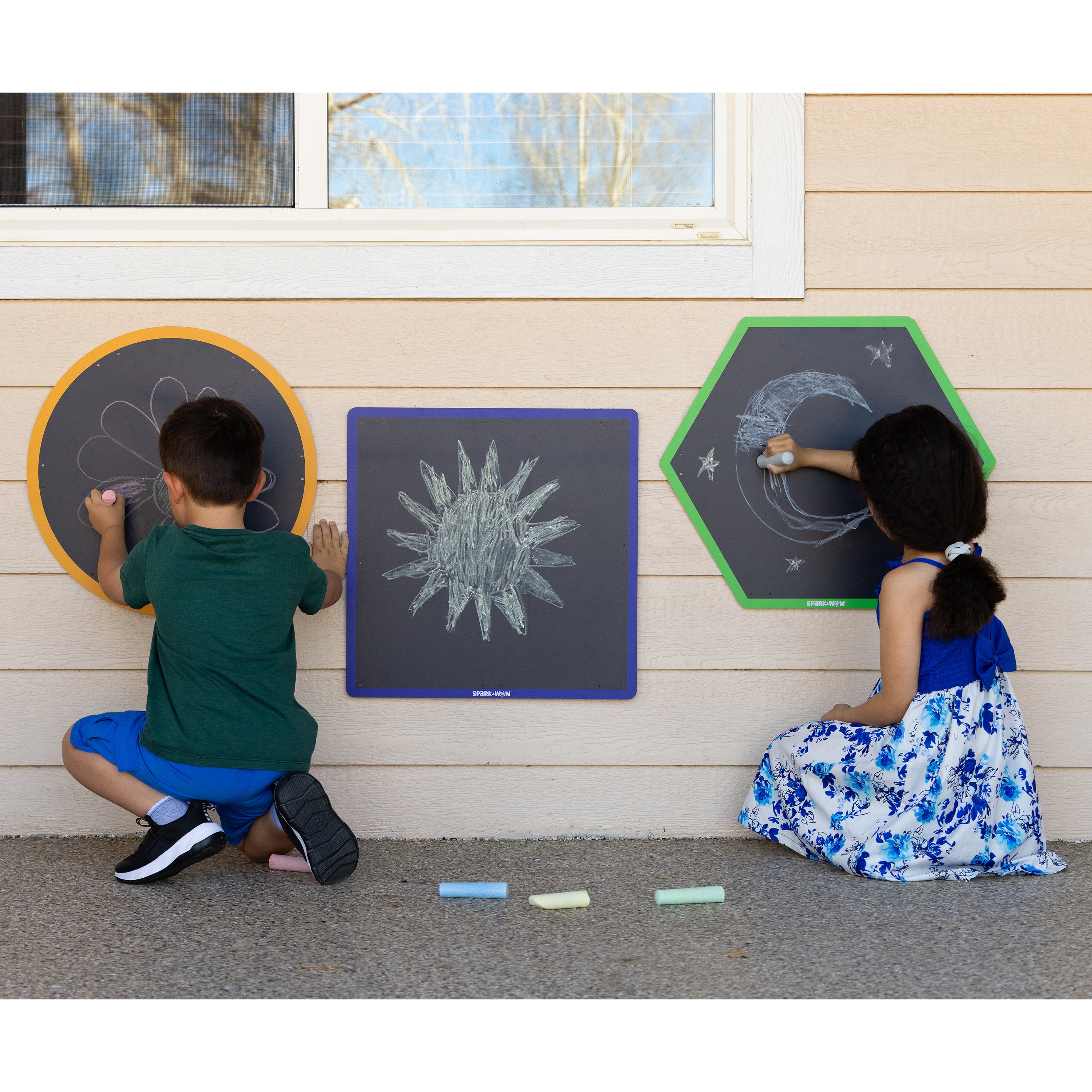 Two children drawing on outdoor chalkboards against a wooden wall.