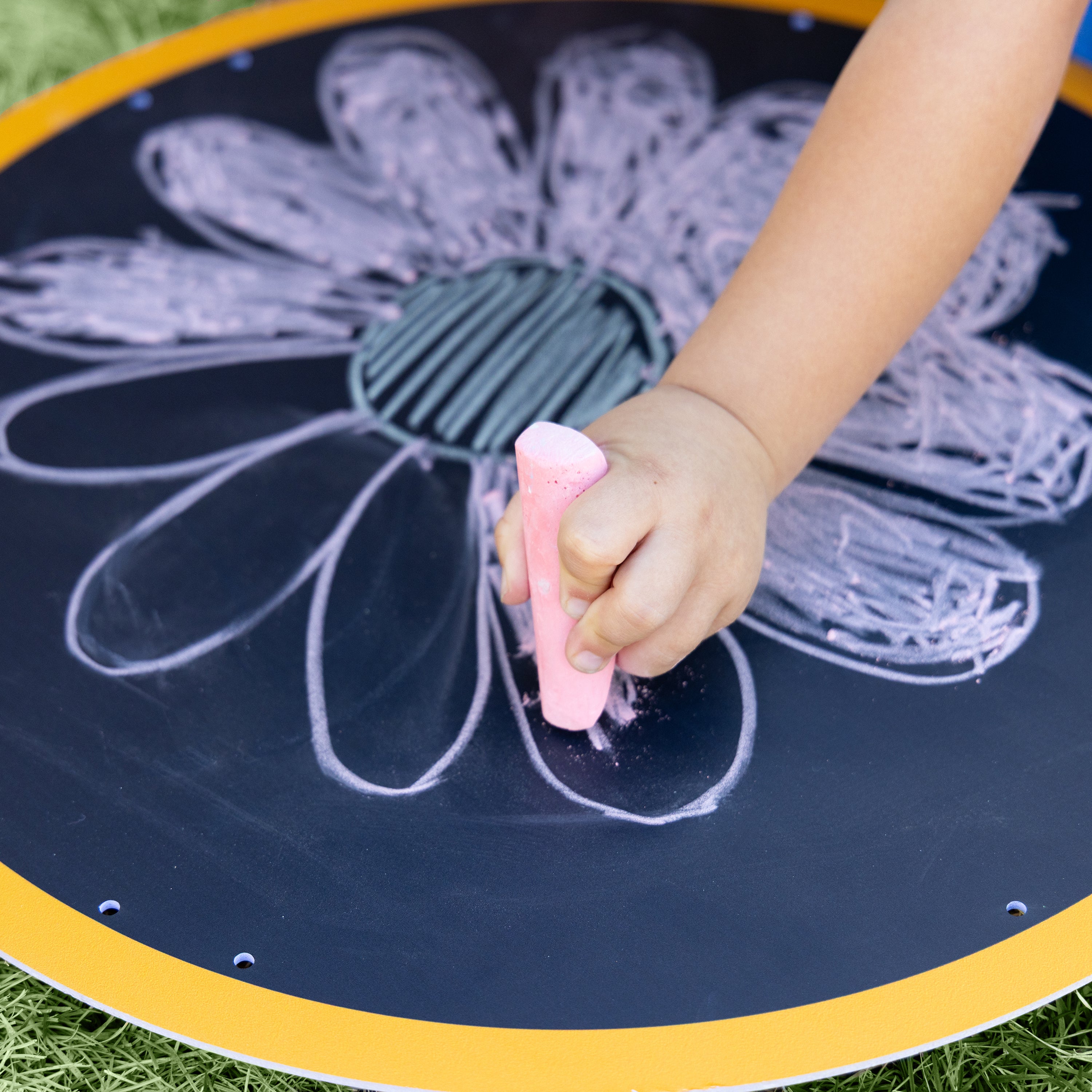 Child's hand holding a pink chalk drawing a flower on a blackboard outdoors.