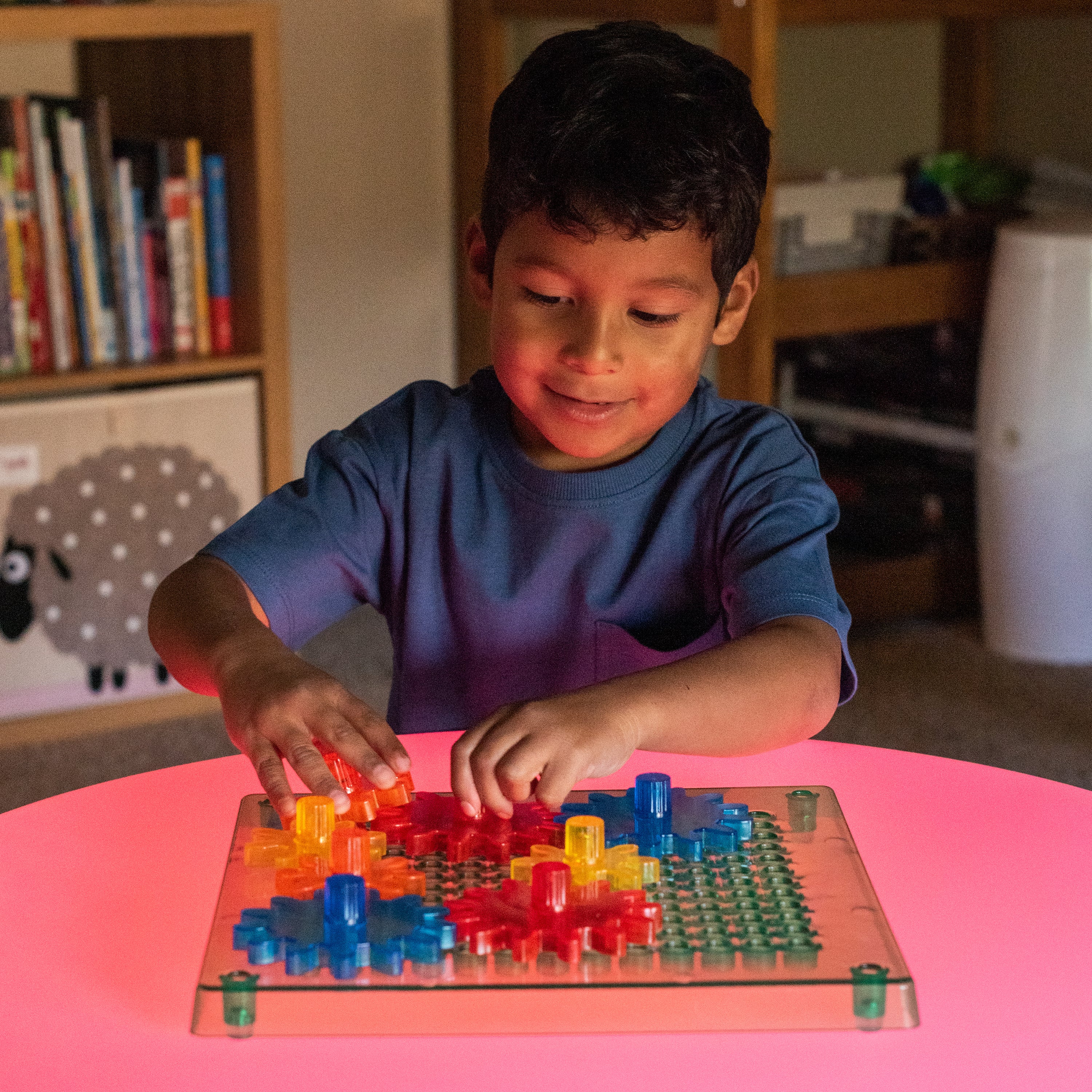 Child playing with a colorful puzzle on a light table in a room with books and a sheep decoration.