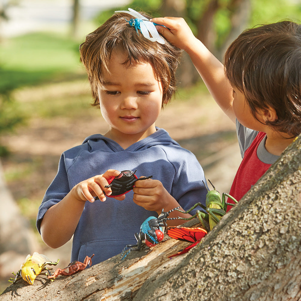 Two children playing with toy insects on a log outdoors.