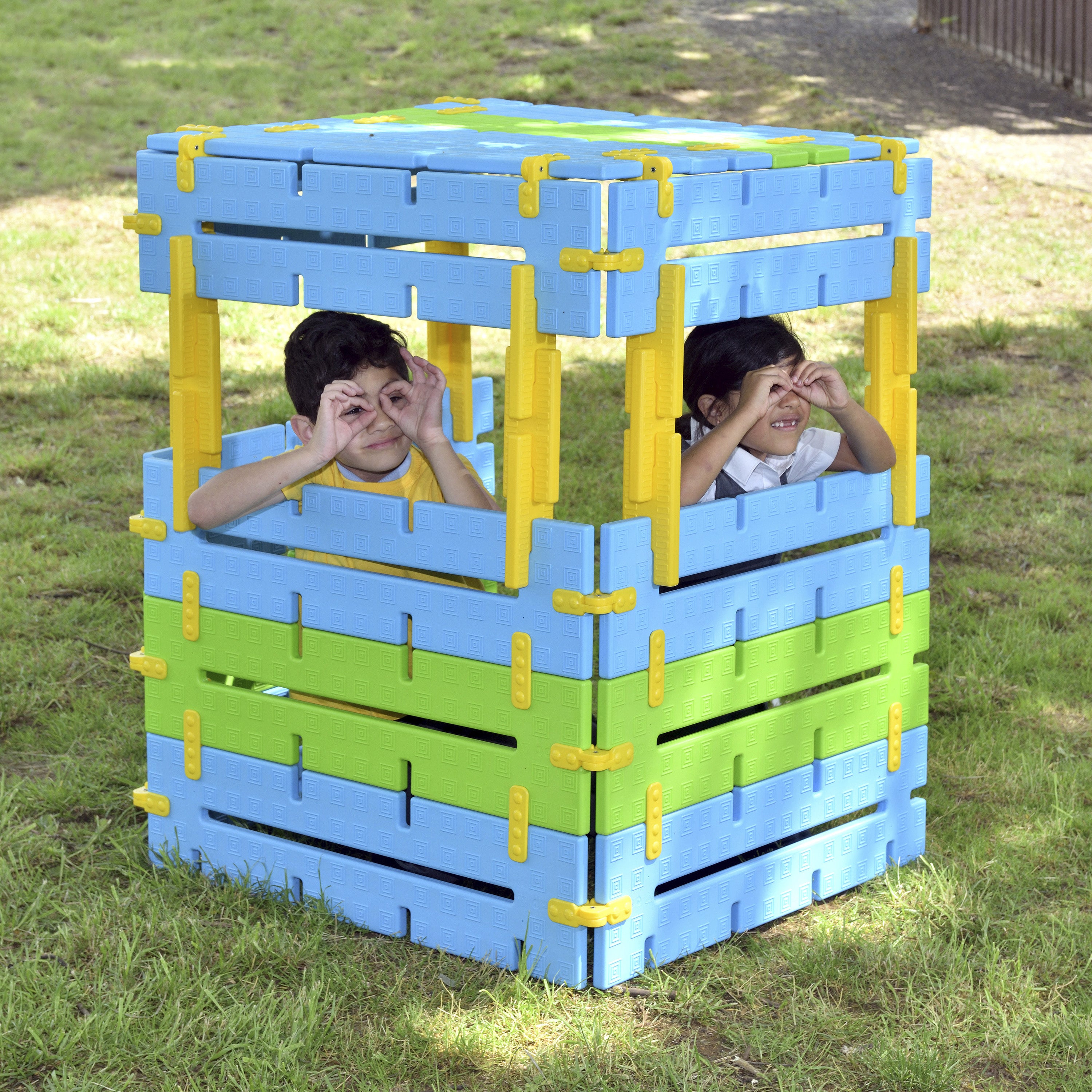 Children playing inside a colorful playhouse made of interlocking plastic bricks on grass.