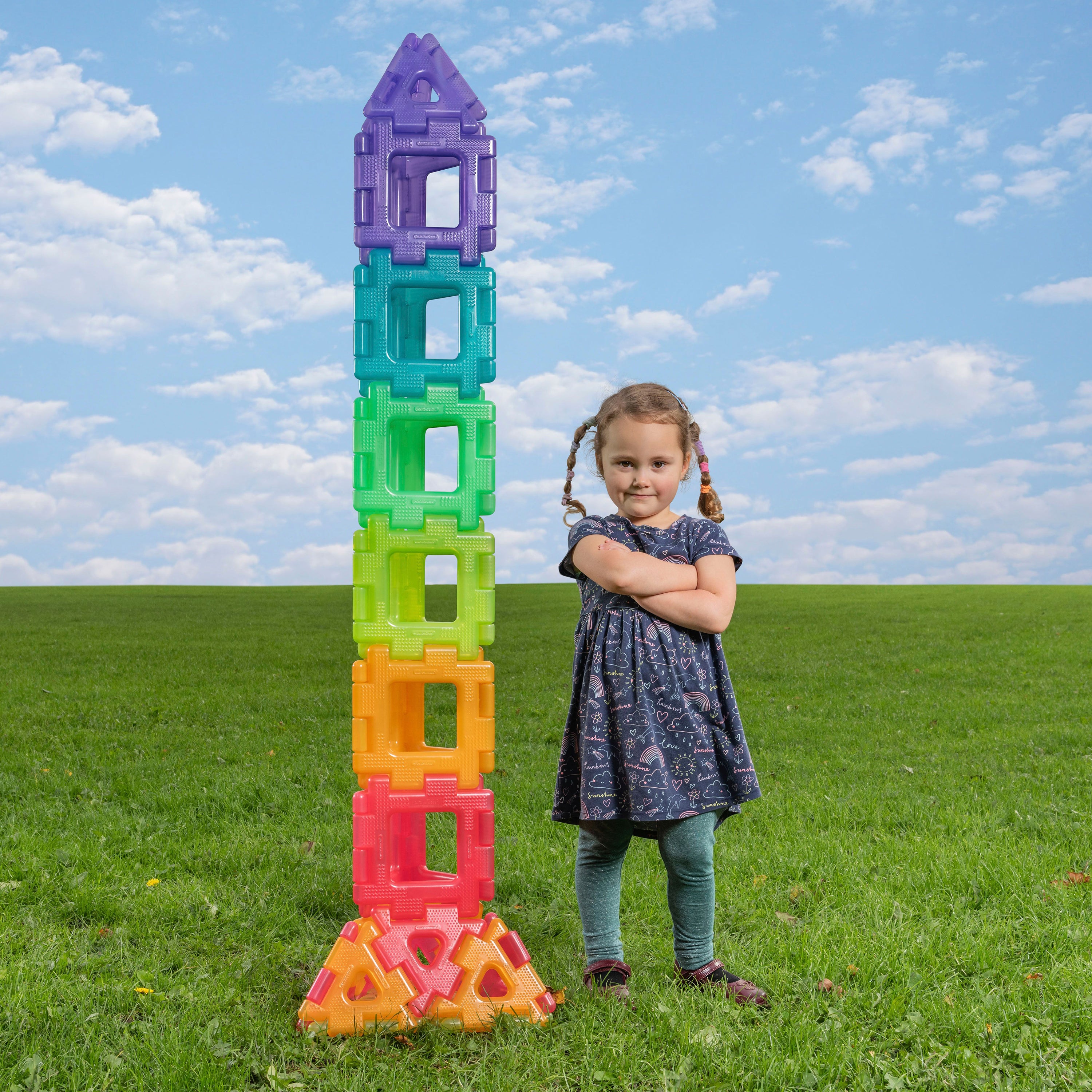 Child standing next to a colorful tower of building blocks on a grassy field with a blue sky.