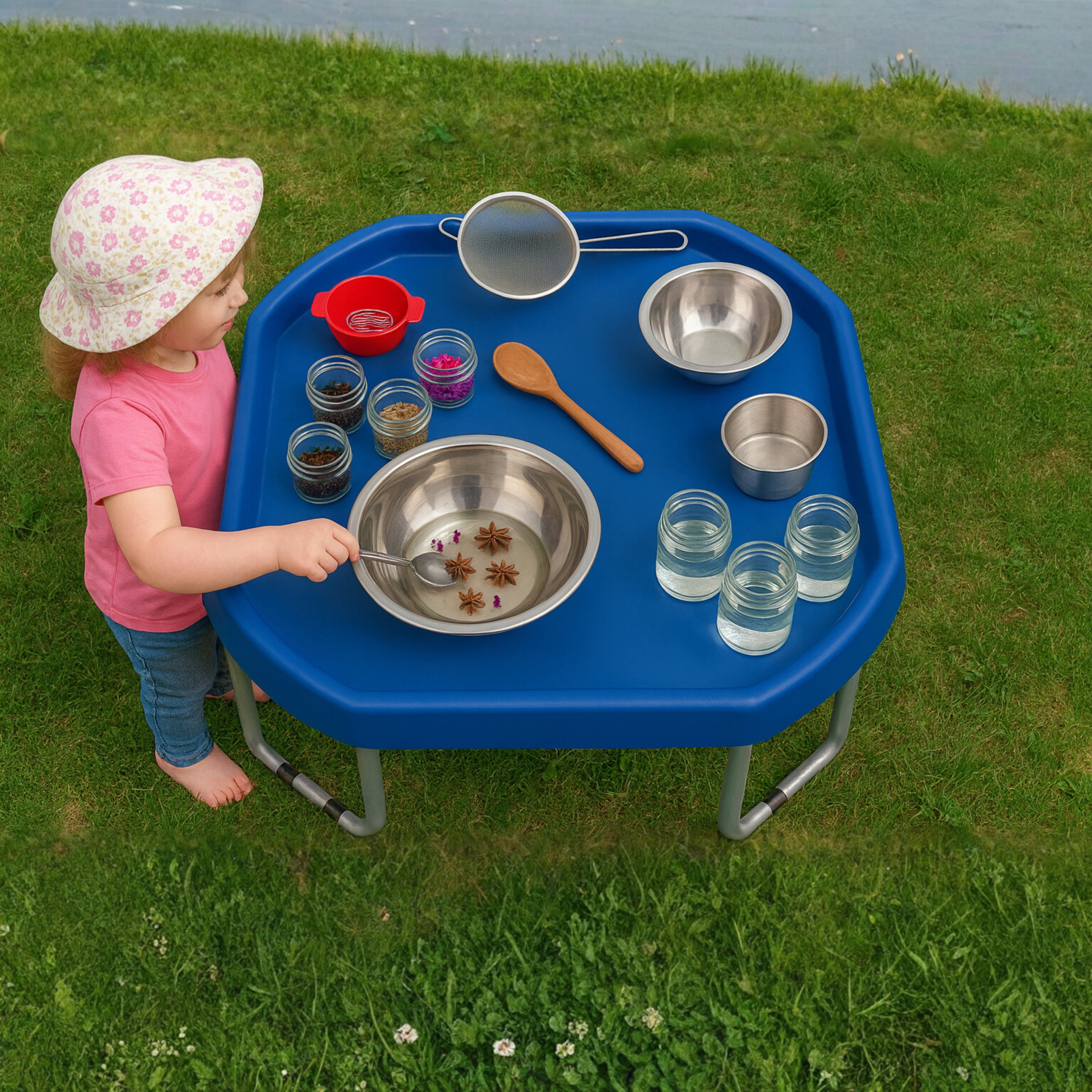 Child playing with a blue outdoor activity table filled with various containers and utensils on a grassy area.