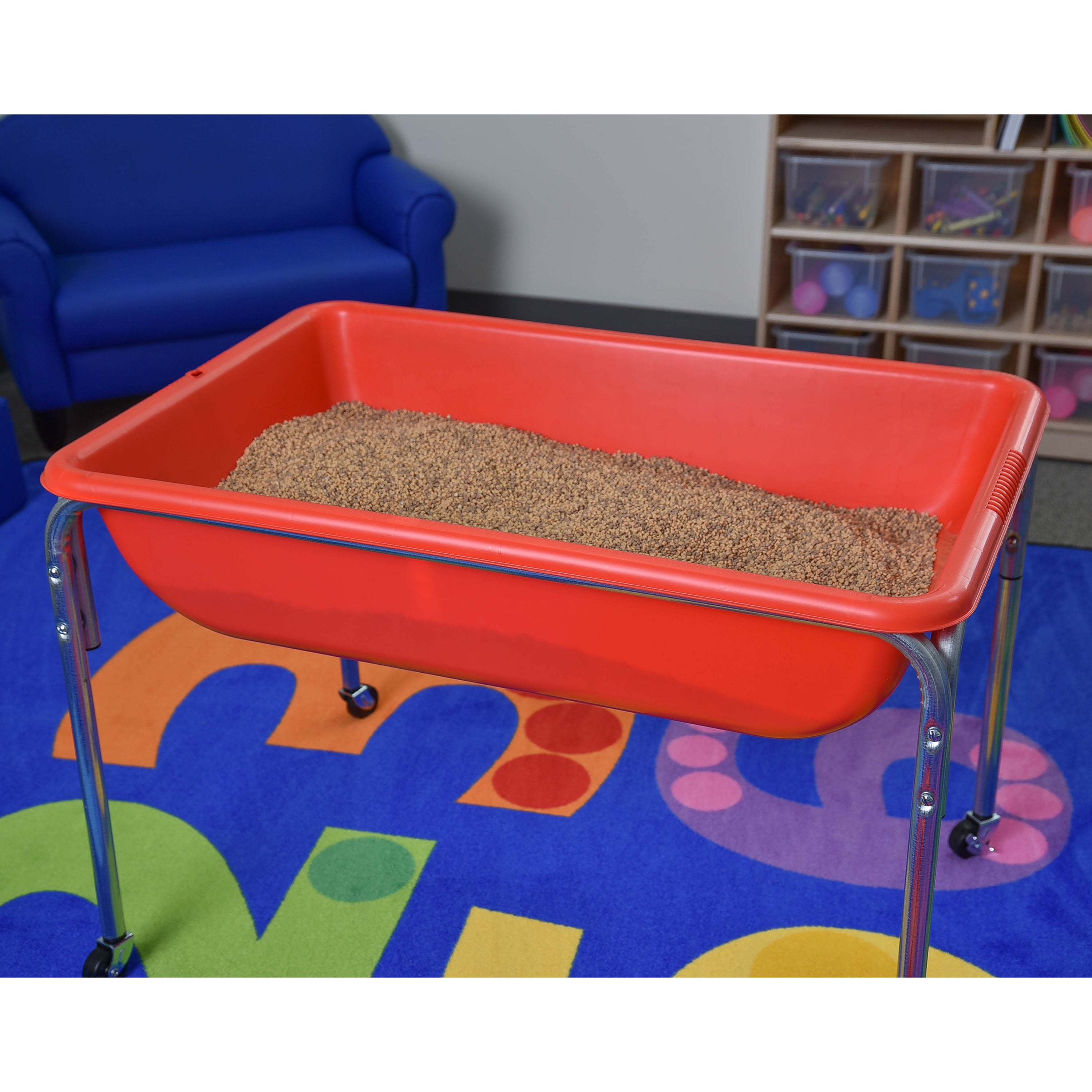 Red sand tray with sand on a colorful floor mat in a room with blue couch and storage shelves.