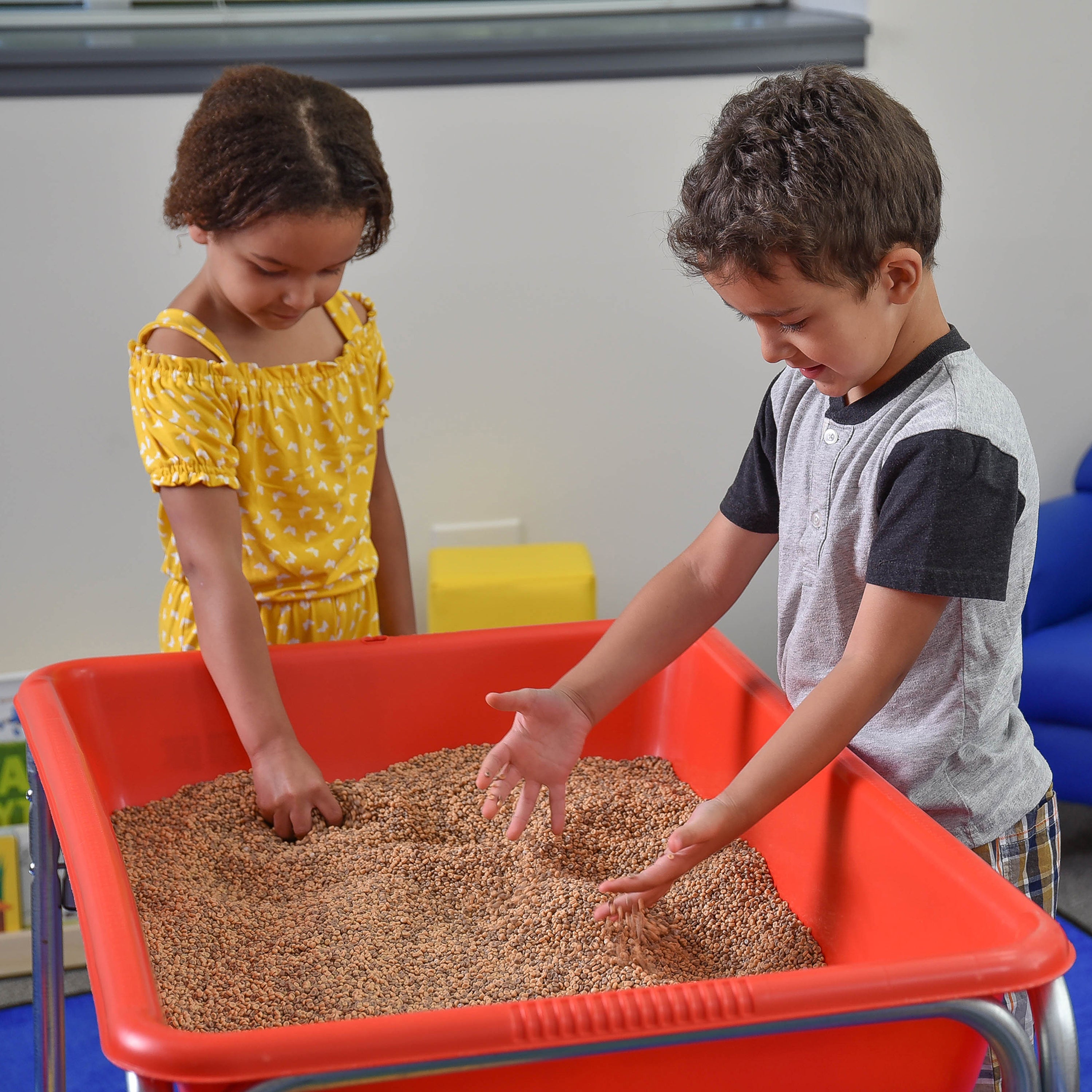 Two children playing with sand in a red tray indoors.
