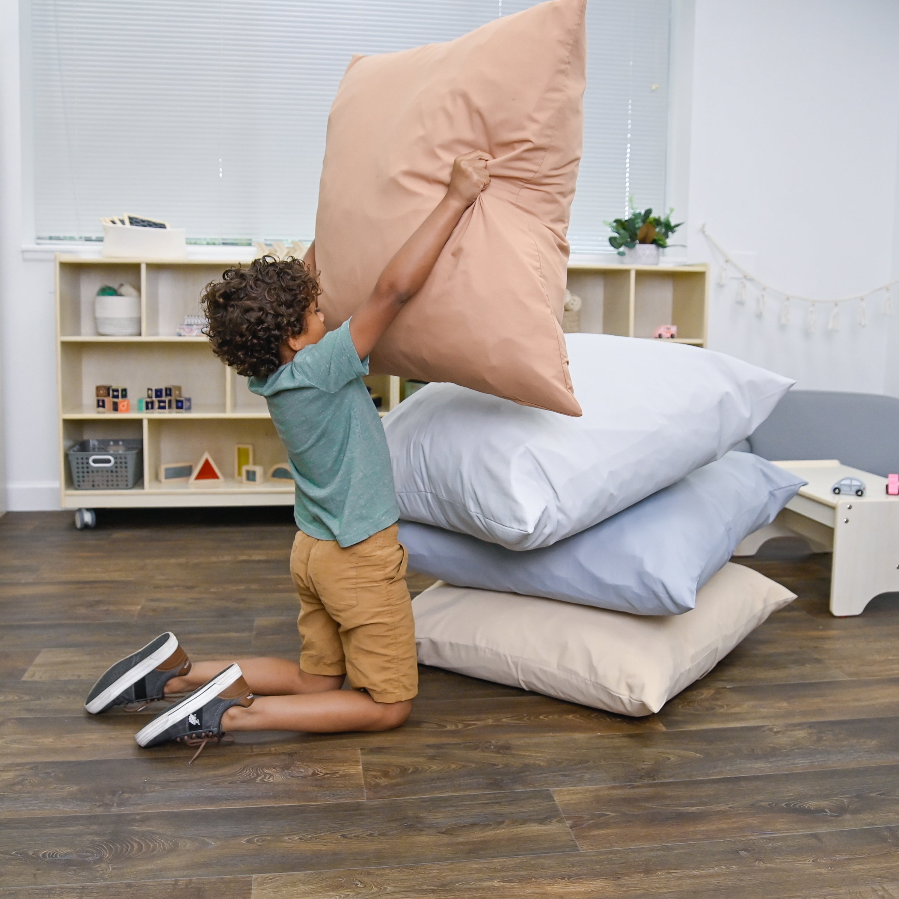 Child playing with pillows on a wooden floor in a room with shelves and a bed.