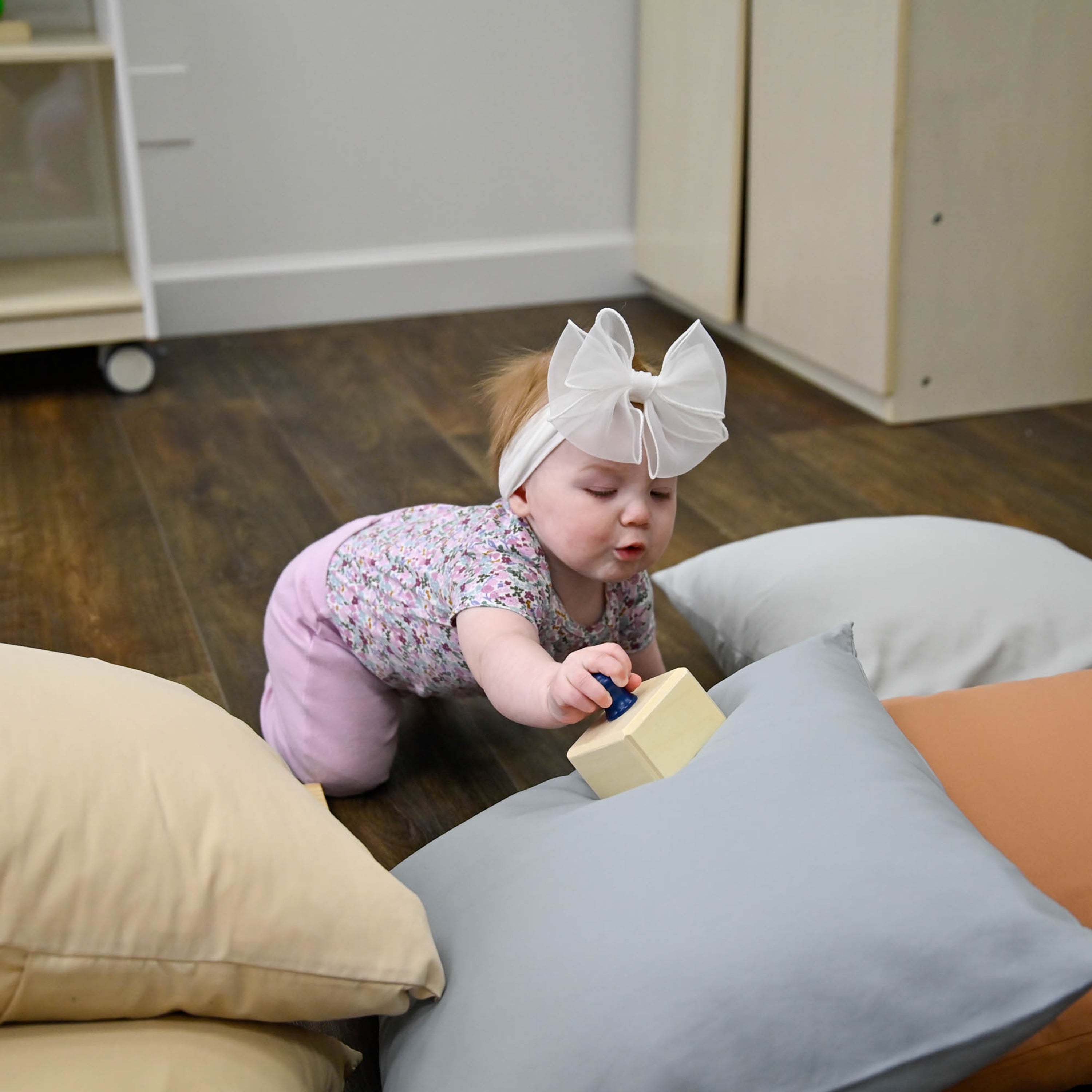 Baby playing with a toy on a wooden floor surrounded by cushions.