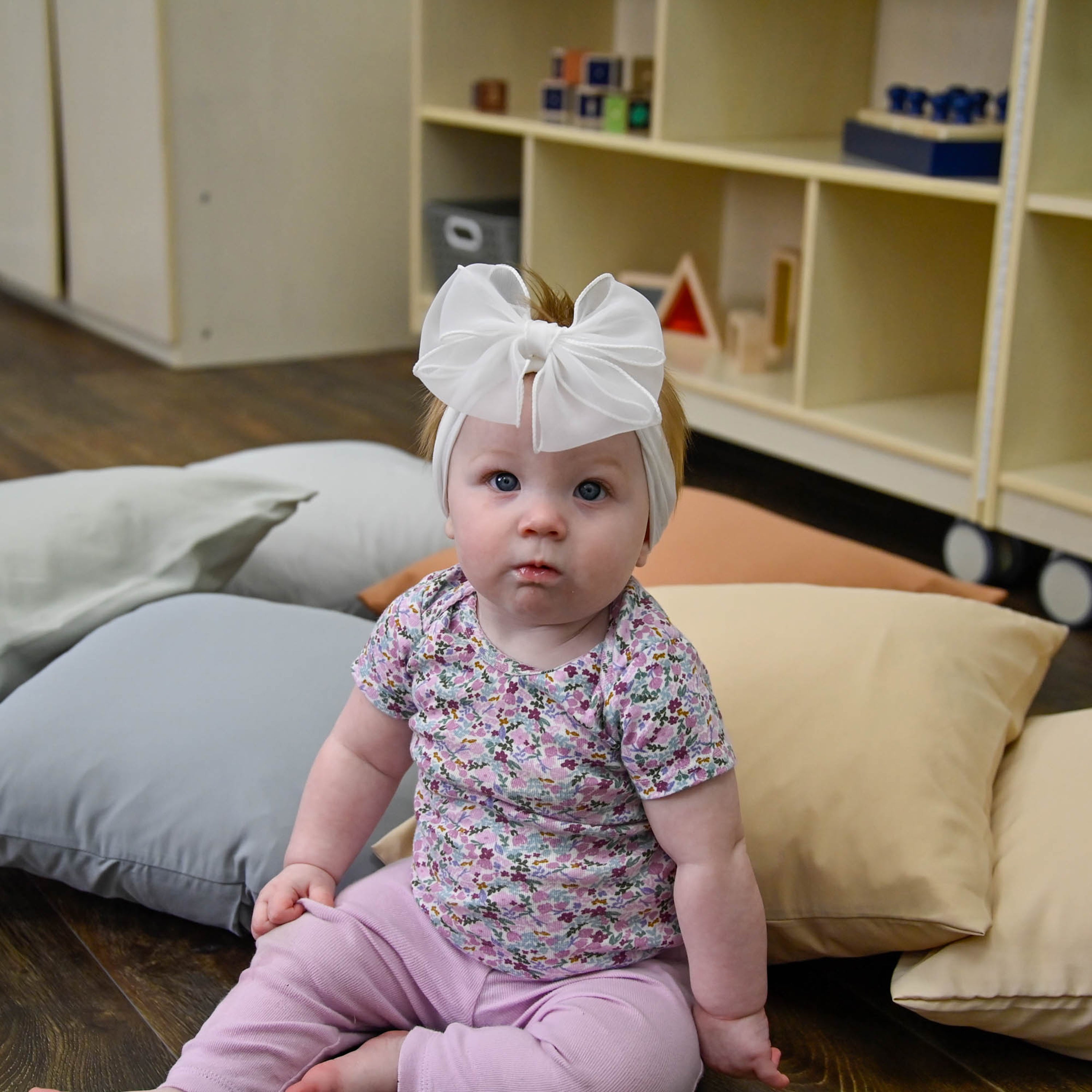 Baby sitting on a couch with colorful cushions in a room with shelves and toys.