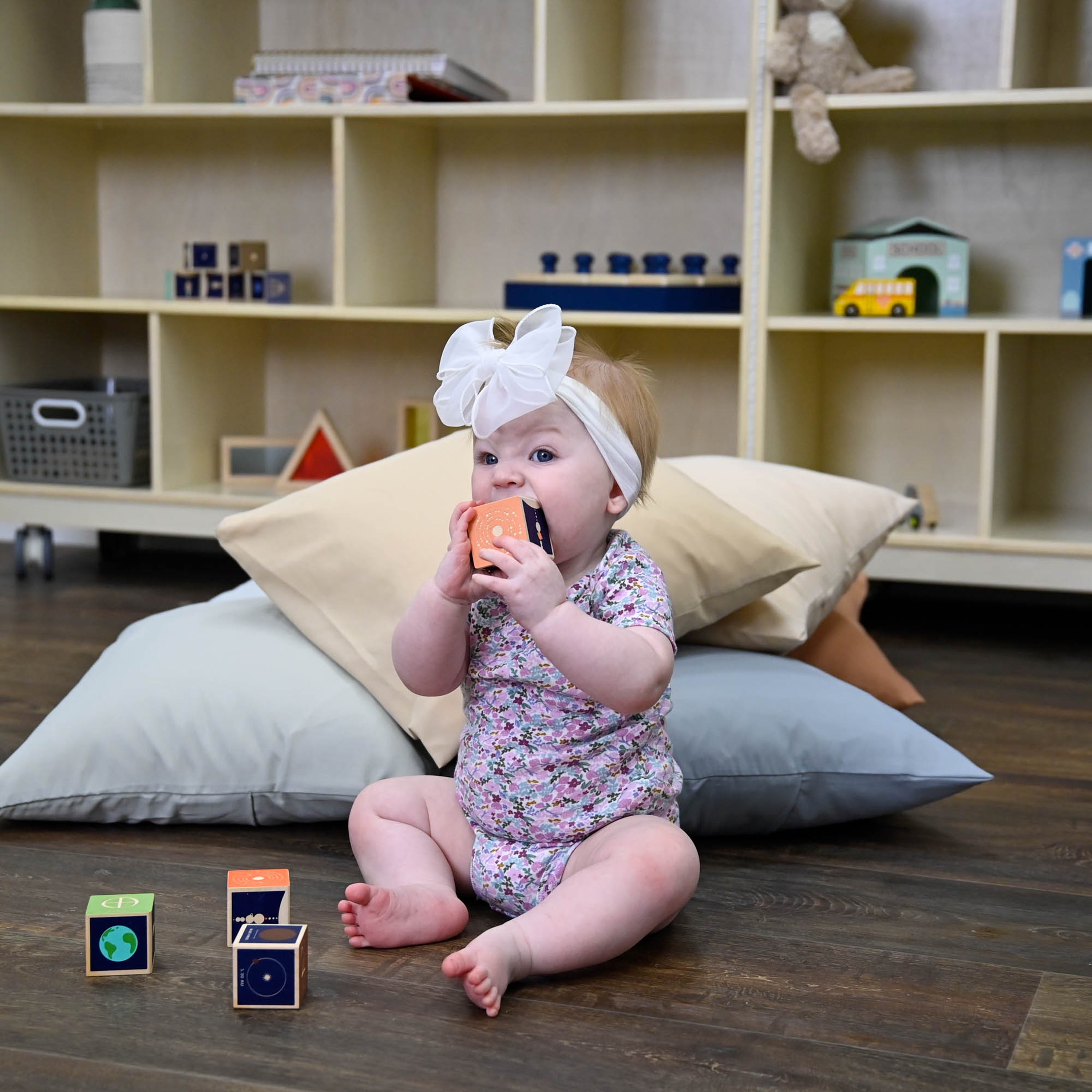 Baby sitting on a wooden floor with toys and pillows, surrounded by shelves with toys in the background.