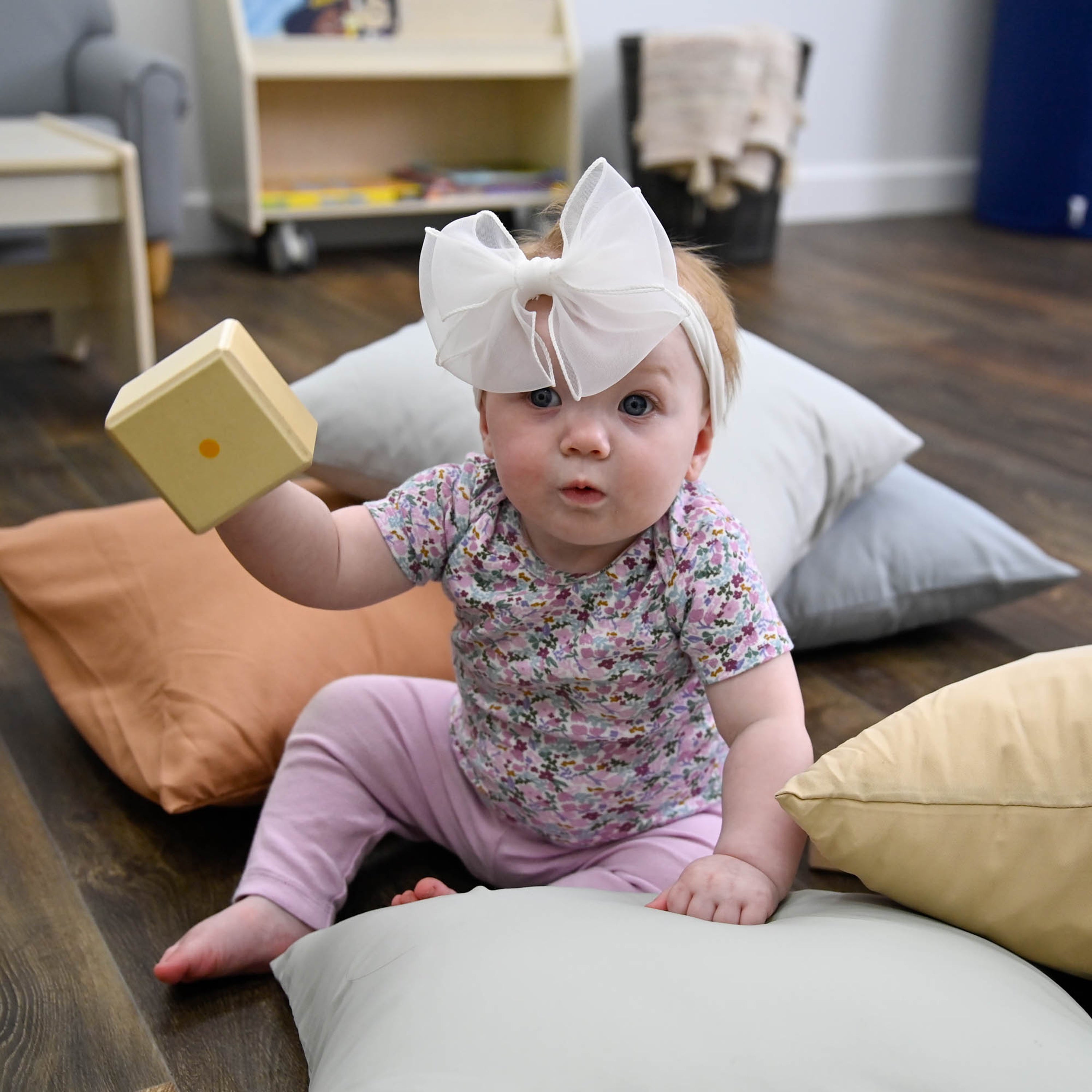 Baby sitting on a floor holding a block with pillows in the background