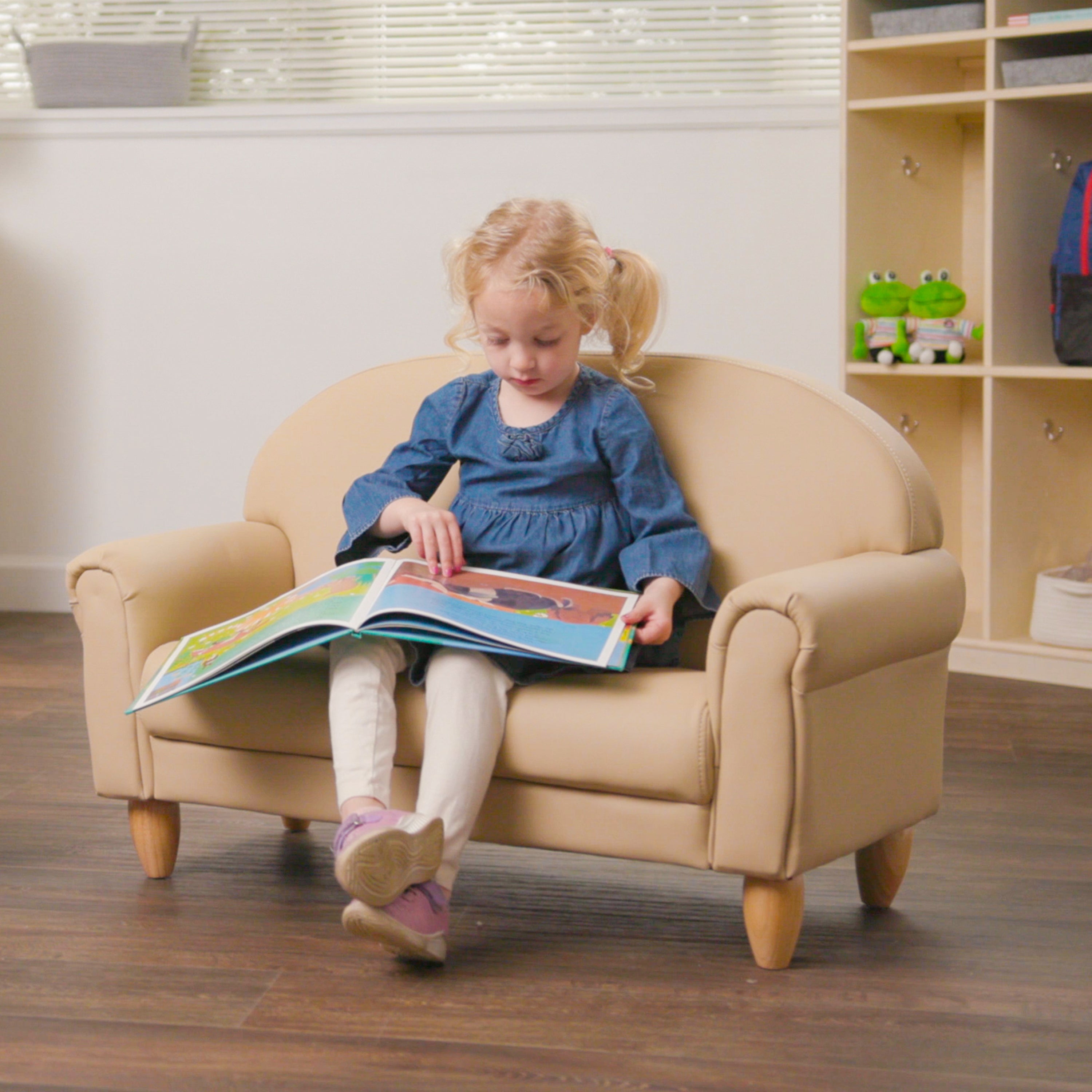 Young girl sitting on a beige chair reading a colorful book in a room with a bookshelf.