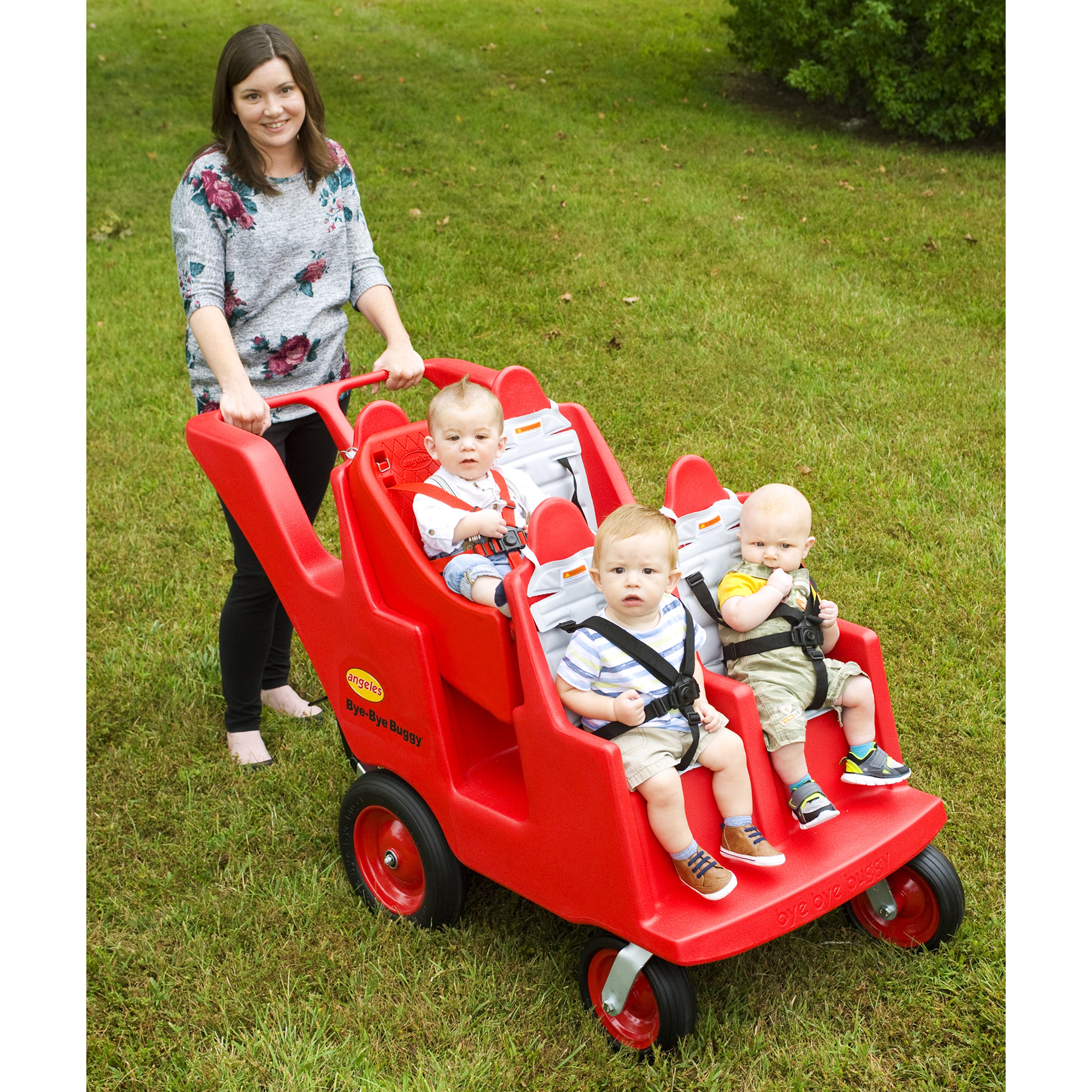 Woman pushing a red four-seater children's cart with three children on a grassy area.