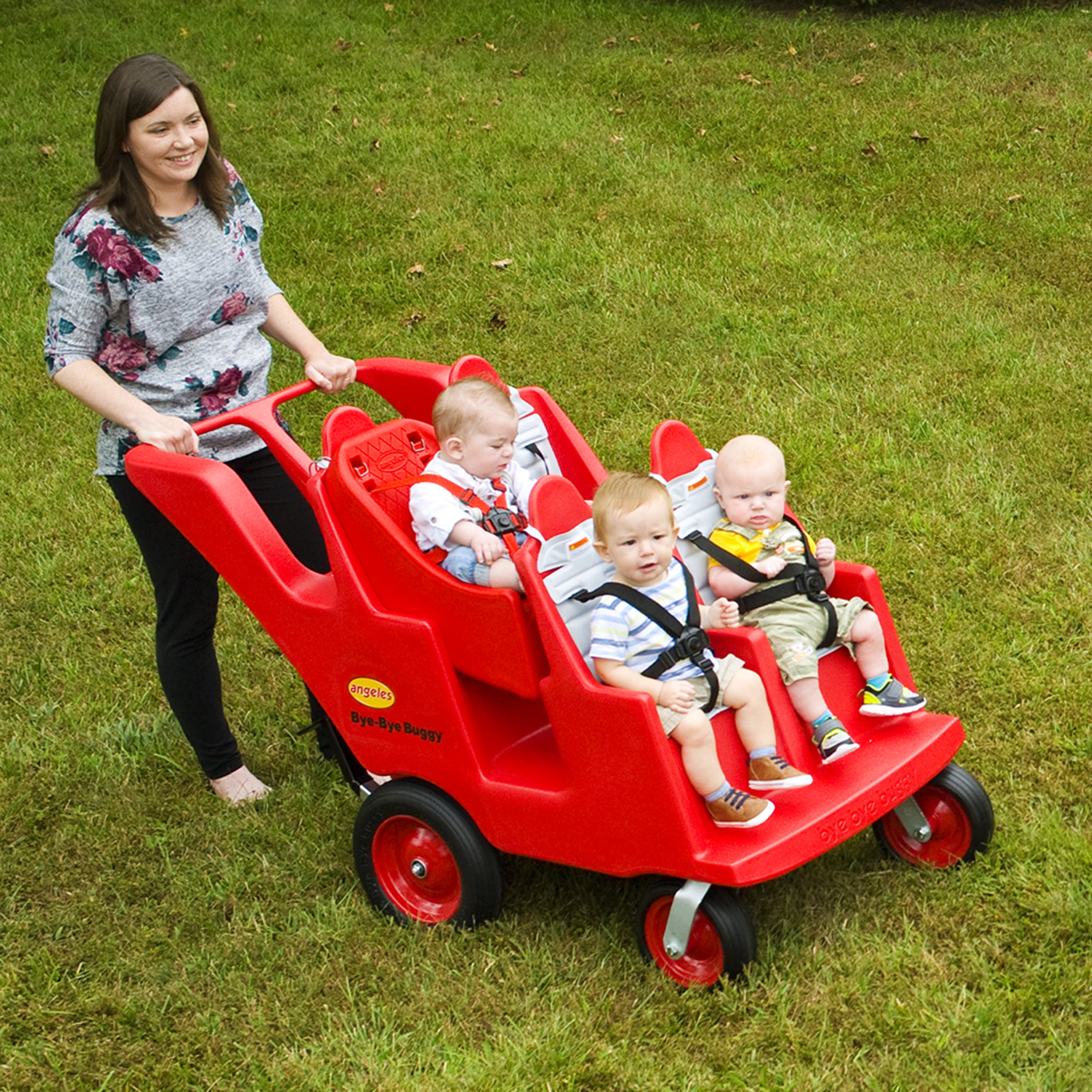 Woman pushing a red four-seater children's wagon with three children on a grassy field.