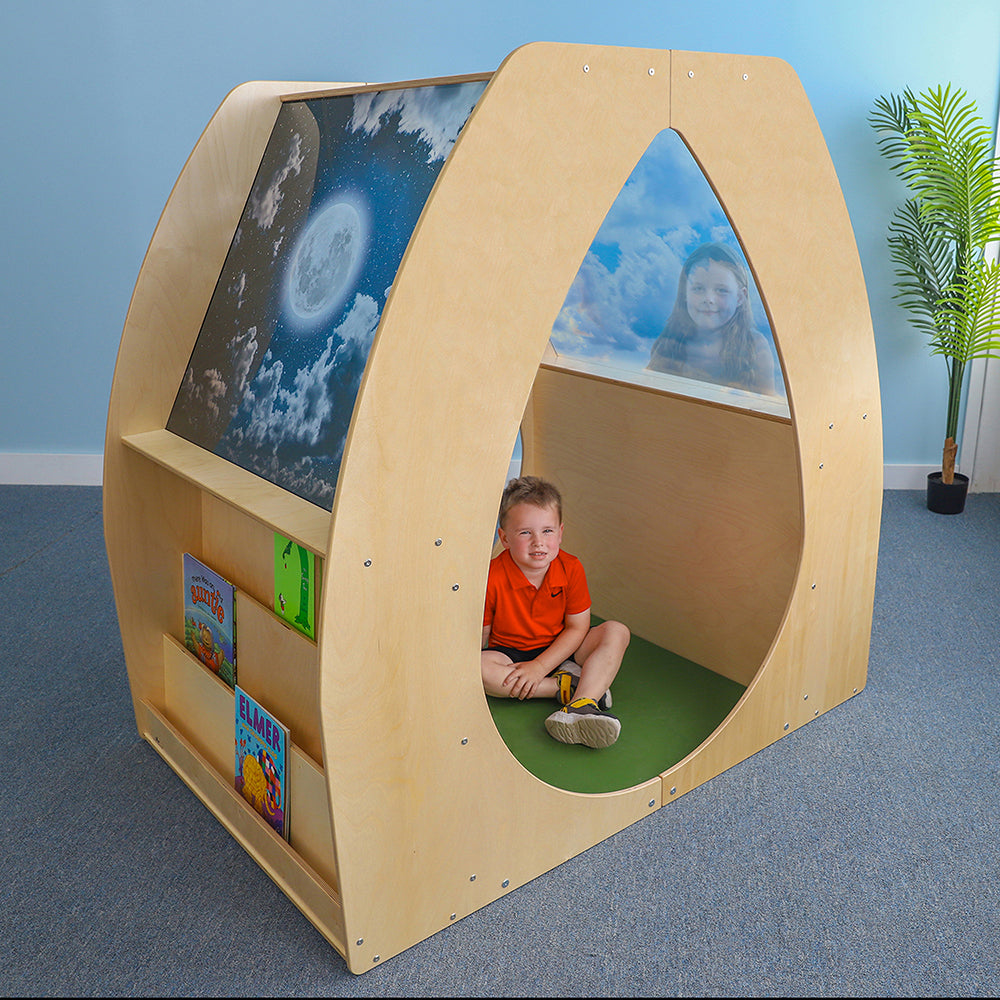 Children's play pod with a child sitting inside, books on shelves, and a plant in the background.