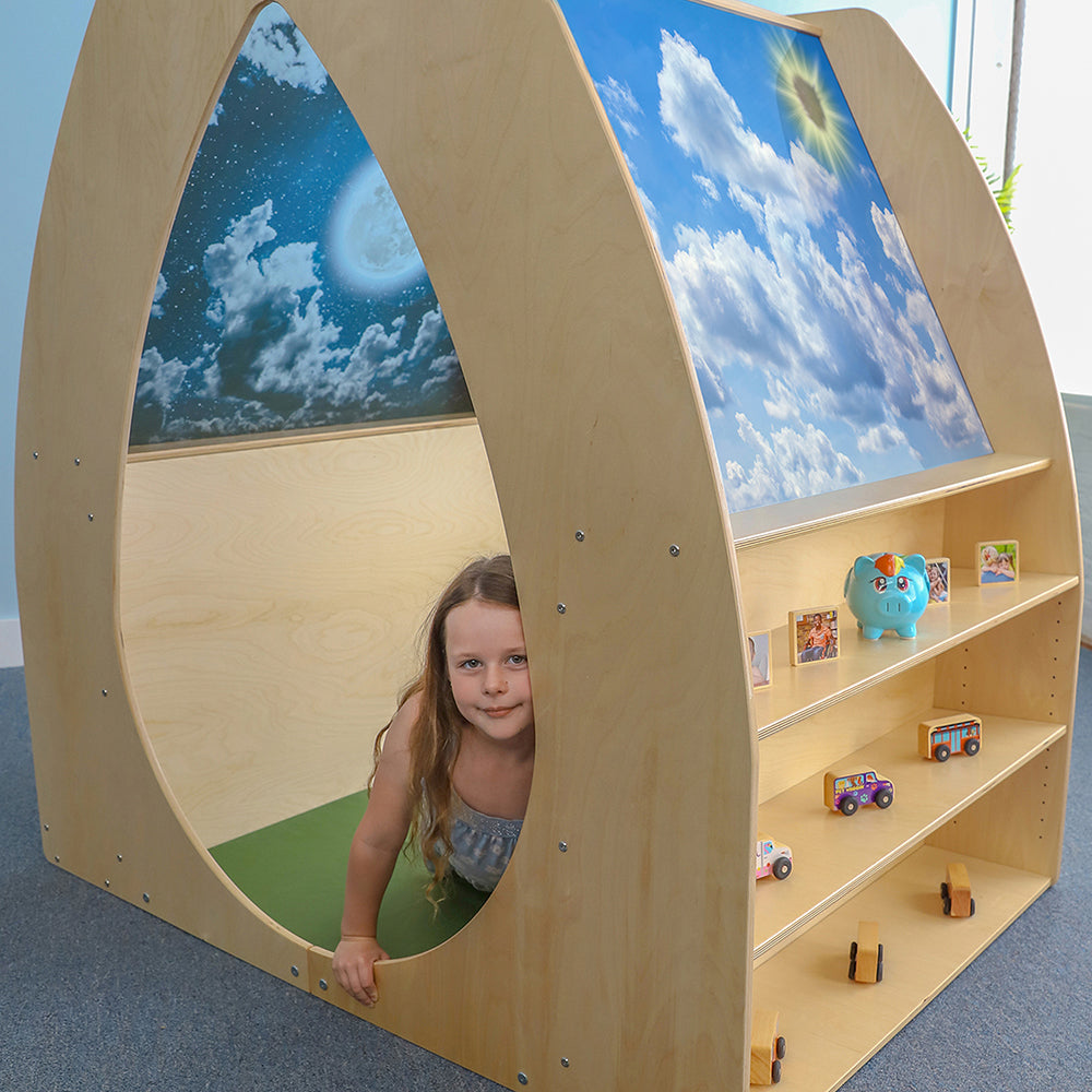 Child playing inside a wooden playhouse with a sky mural, shelves with toys, and a piggy bank.