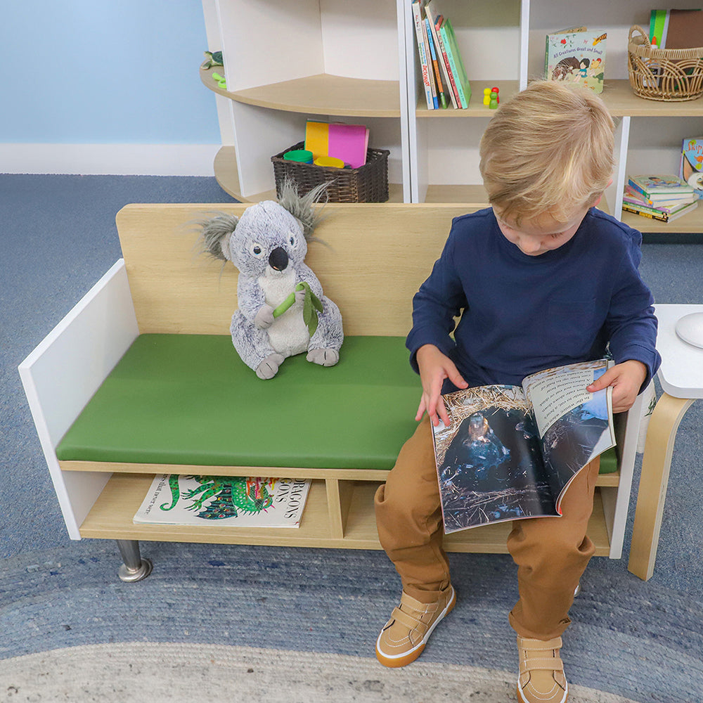 Child reading a book on a bench with a stuffed koala toy in a room with shelves and books.