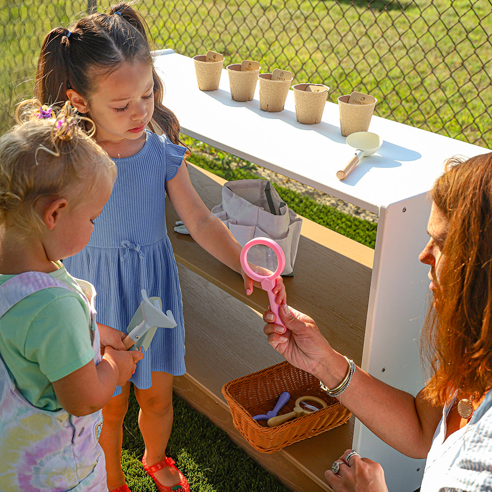 Two young girls interacting with a woman outdoors, possibly at a play area or educational setting.